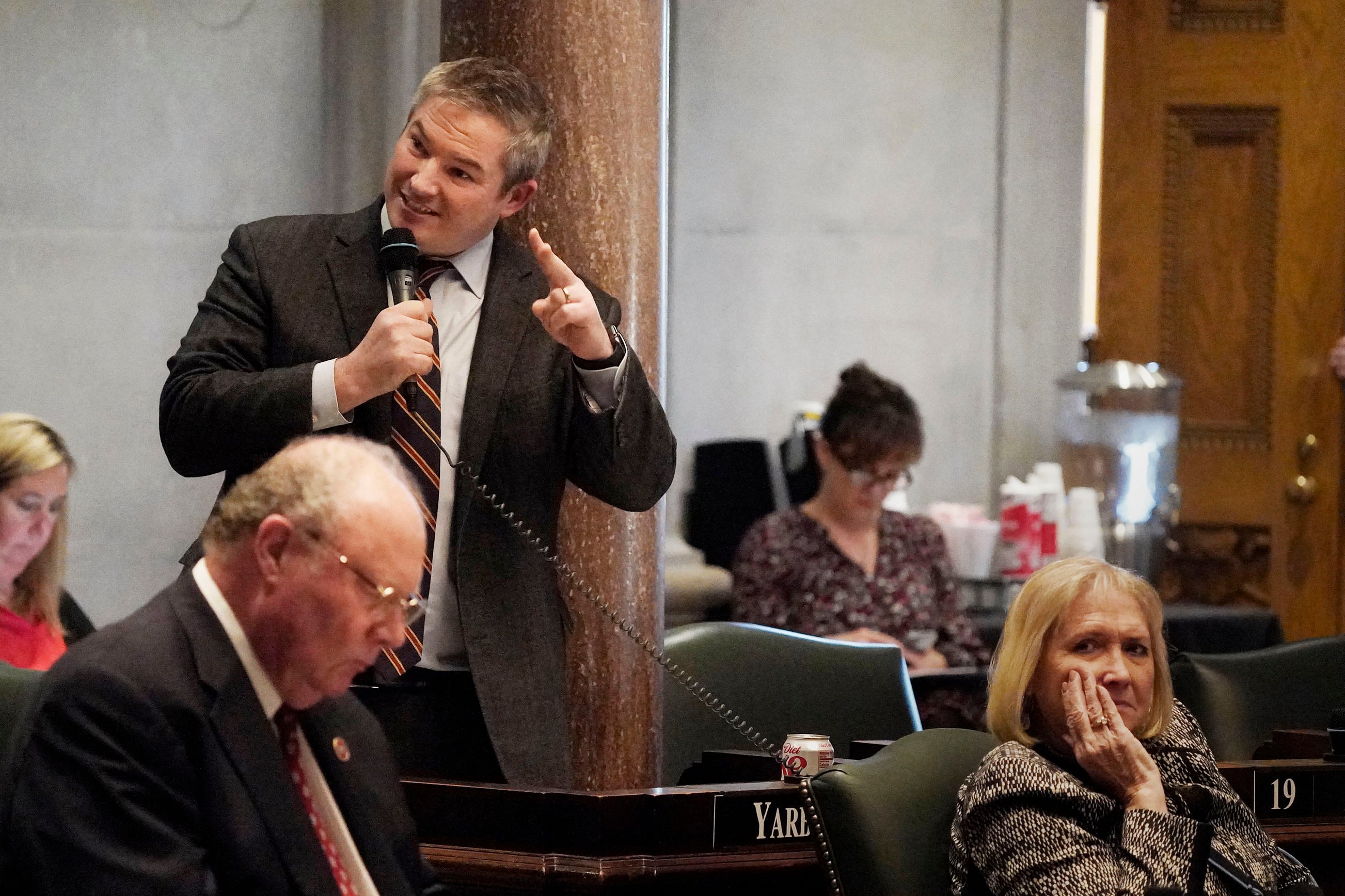 A man wearing a suit and standing in front of a desk speaks into a microphone in a legislative chamber.