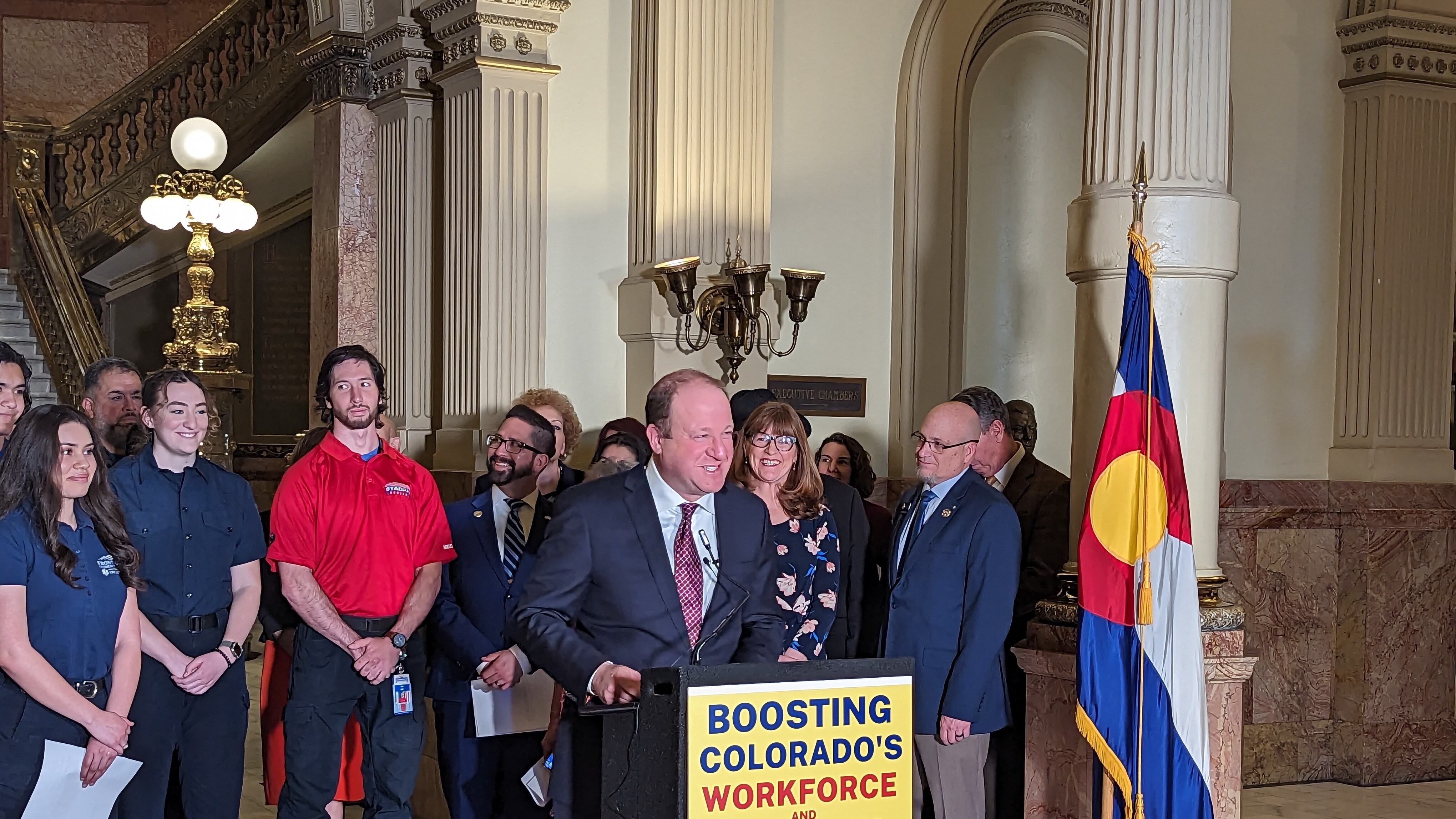 Colorado’s governor in a blue suit stands at a podium with a yellow sign surrounded by people.