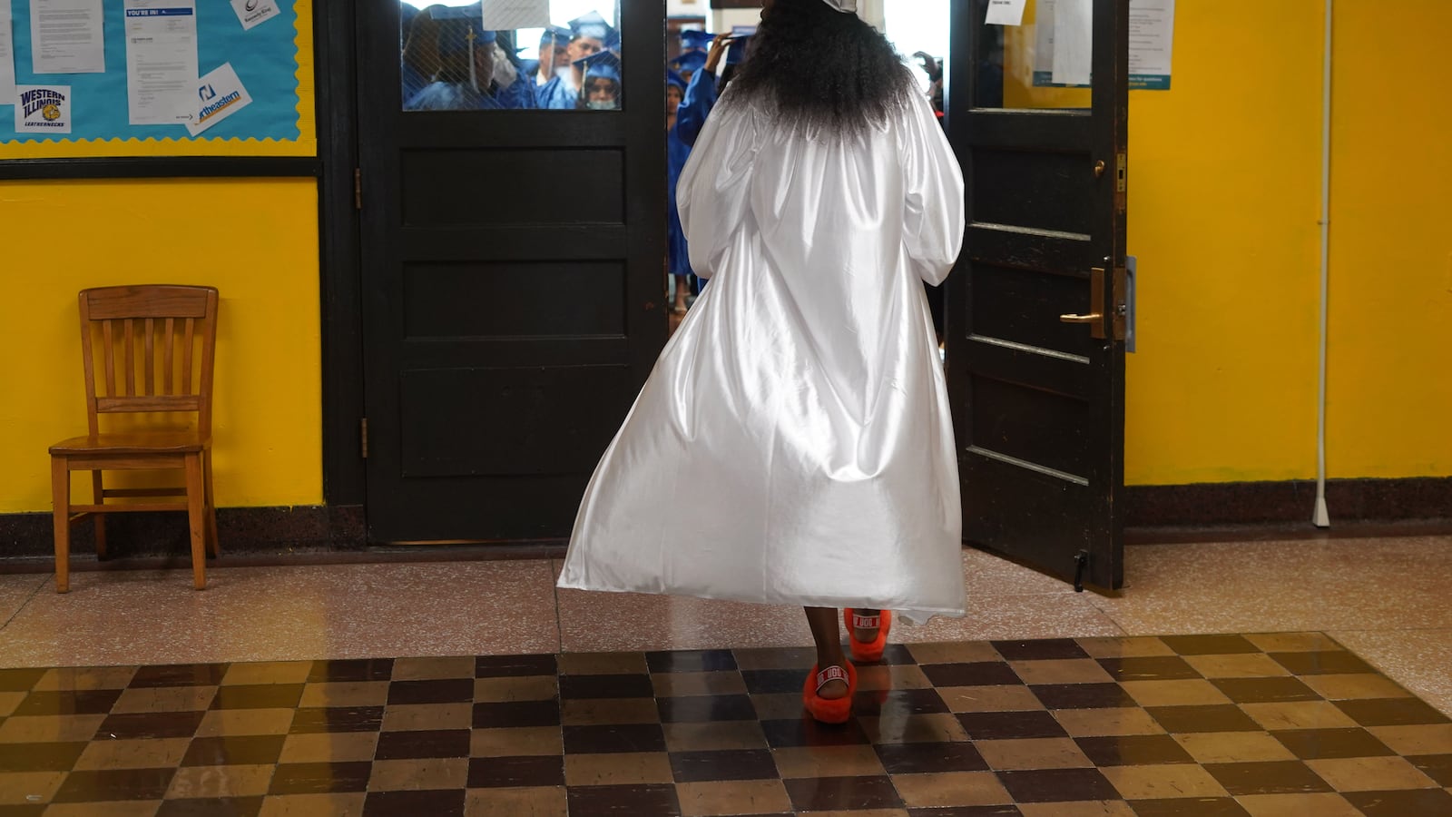 A student walks through a doorway in a white graduation cap and gown.