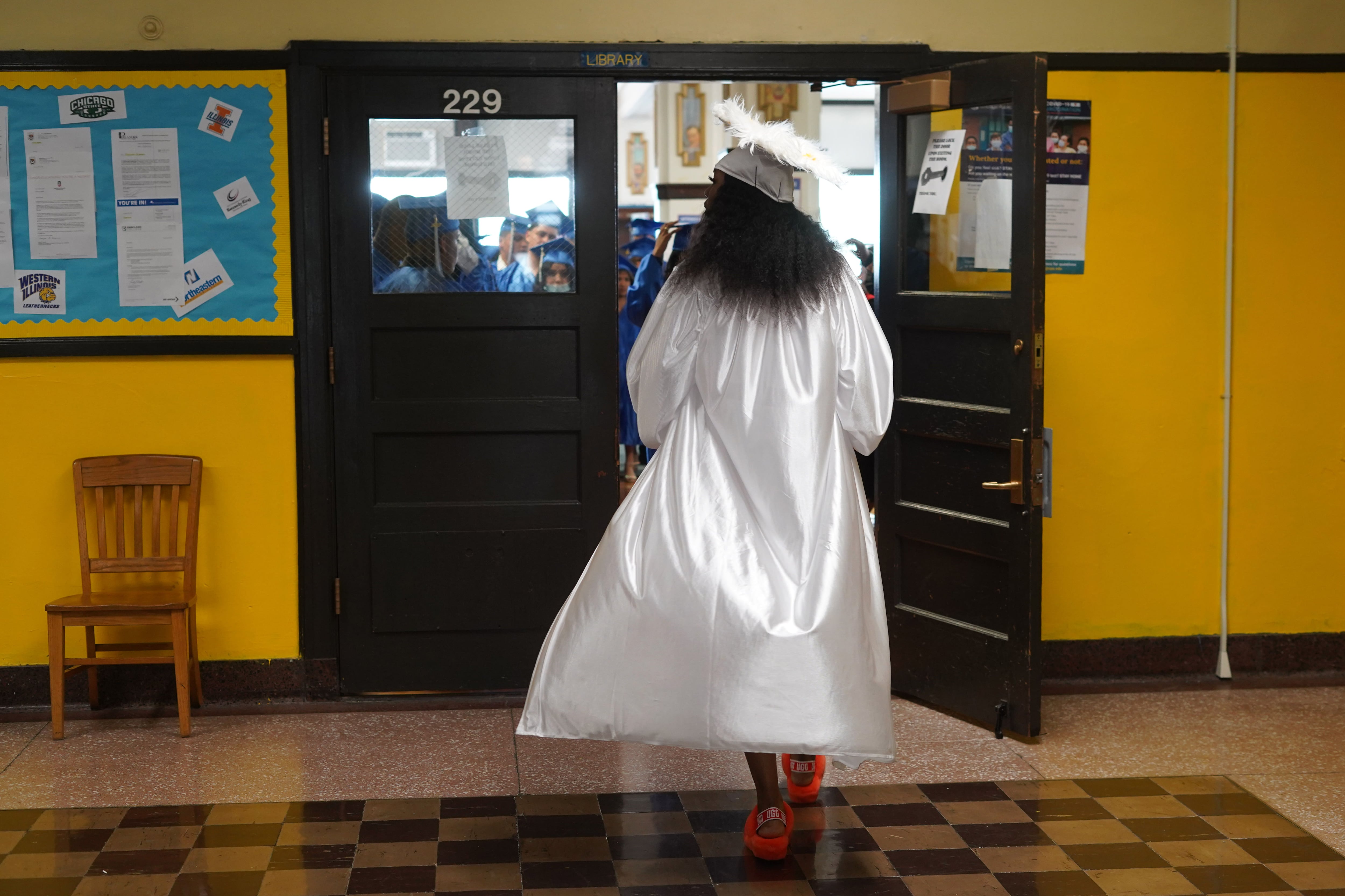 A student walks through a doorway in a white graduation cap and gown.