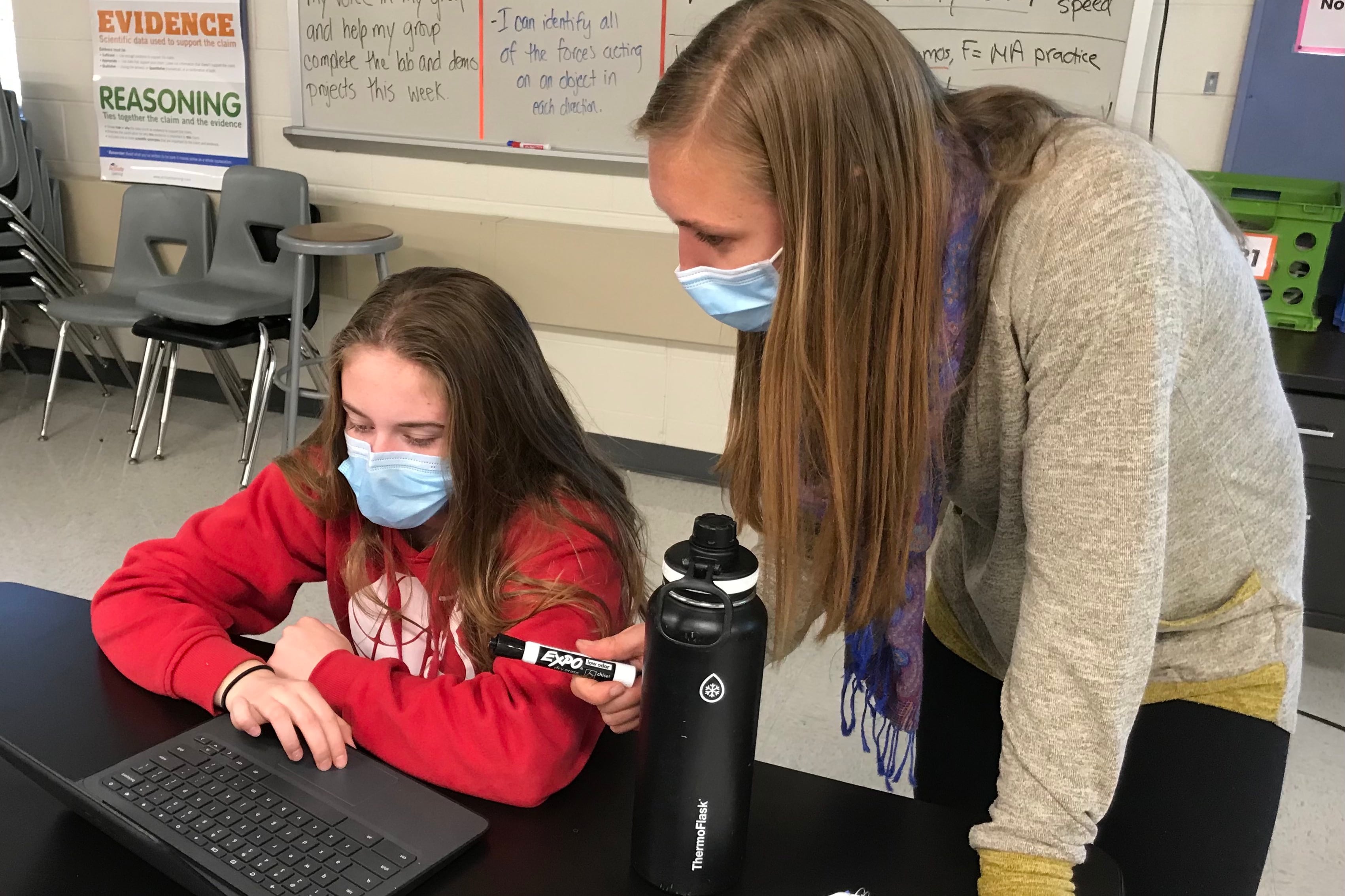 A masked teacher leans over to help a masked middle school student looking at a laptop.