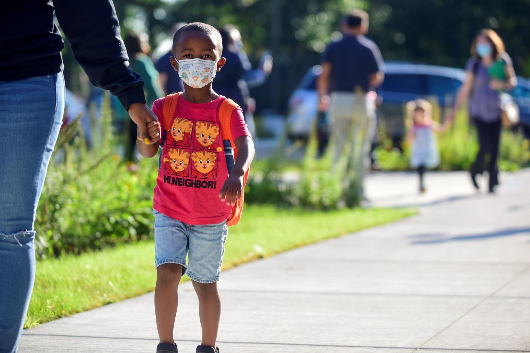 A little boy wearing a red shirt and protective mask holds someone’s hand as other children walk behind them on a sidewalk.