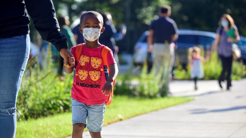A little boy wearing a red shirt and protective mask holds someone’s hand as other children walk behind them on a sidewalk.