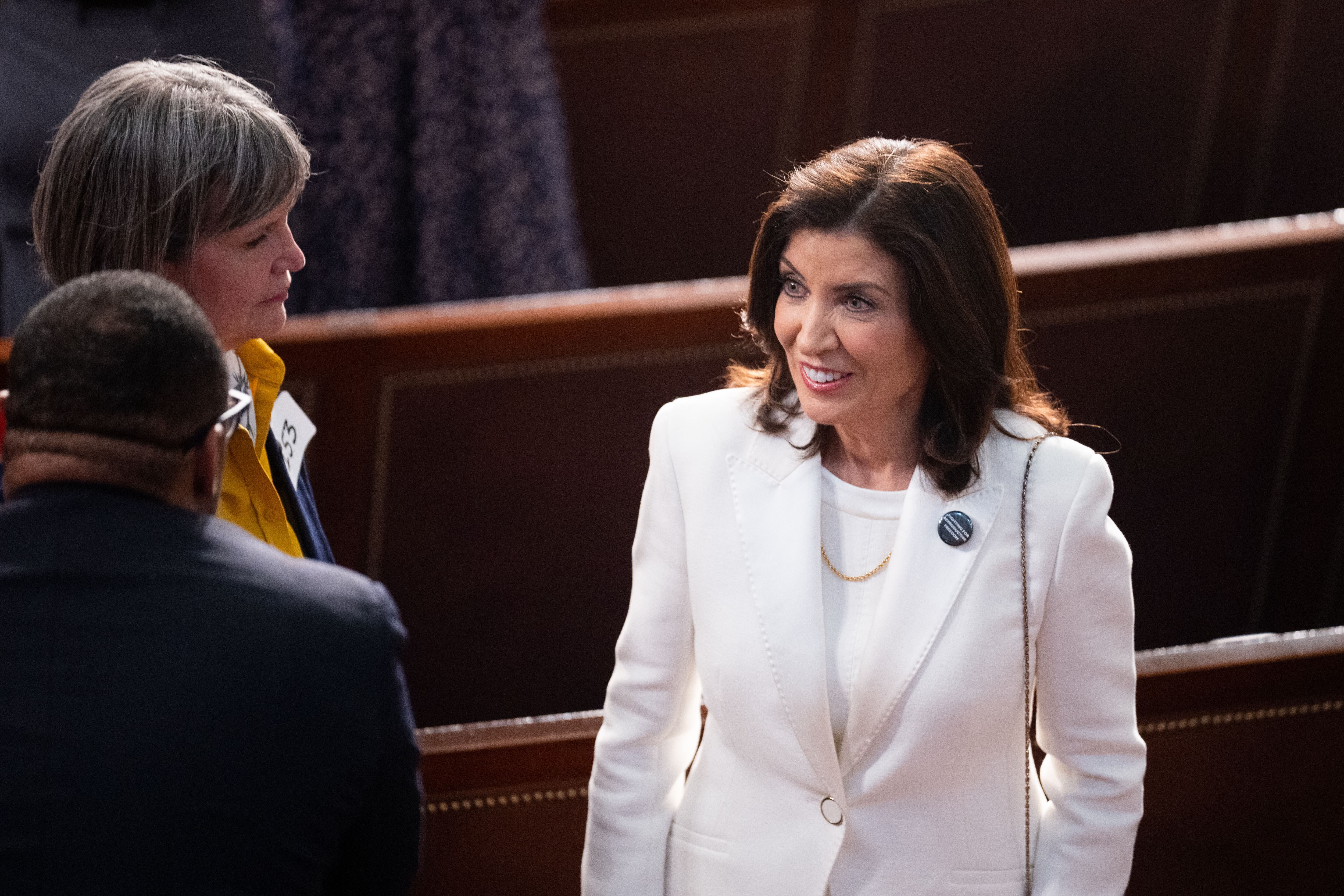 A woman with short dark hair and wearing a white suit stands near two other people with two wooden benches in the background.