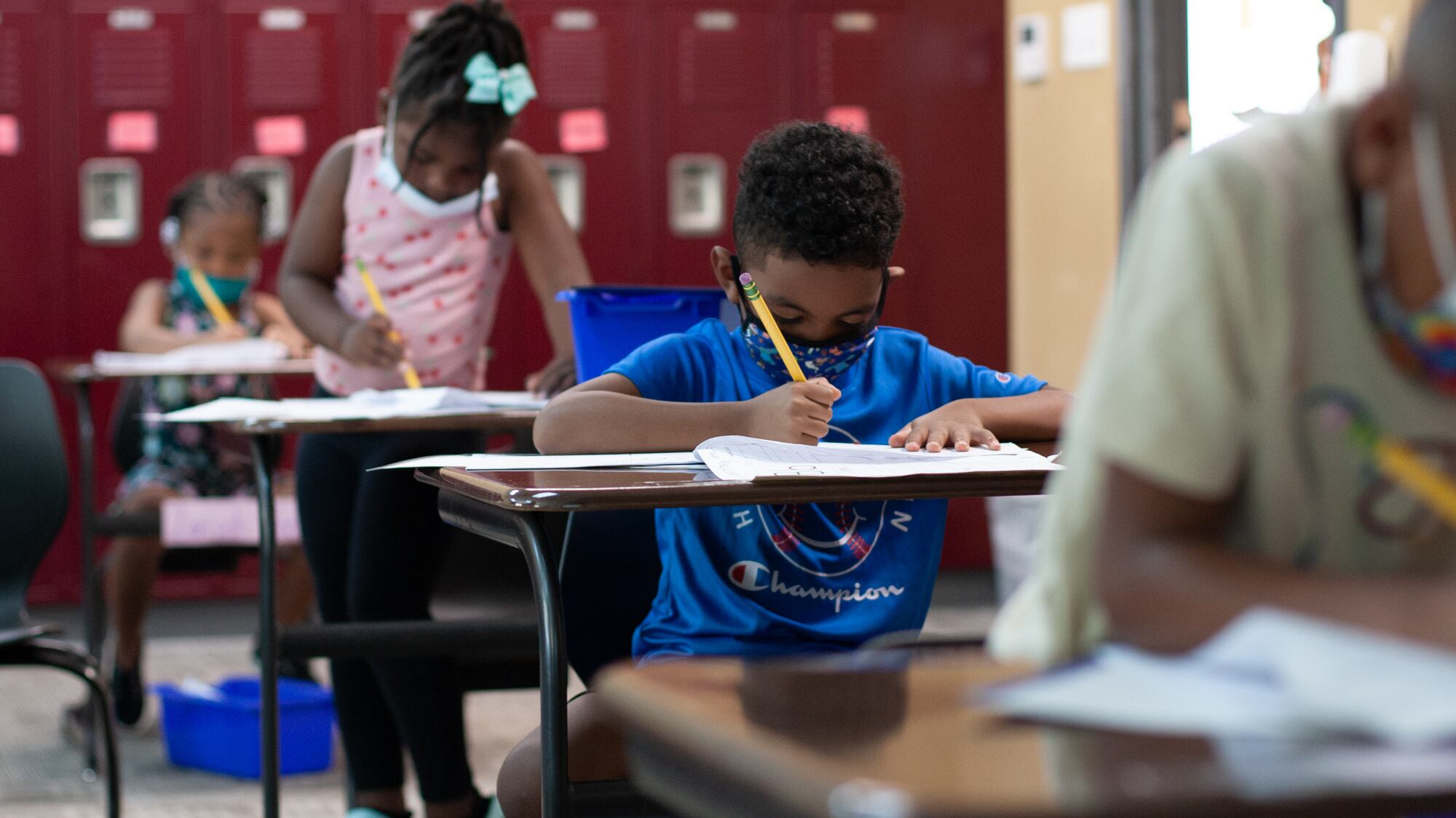 Four students sit in a row of desks in a classroom lined with red lockers. One boy wears a blue shirt and blue mask while writing on a worksheet