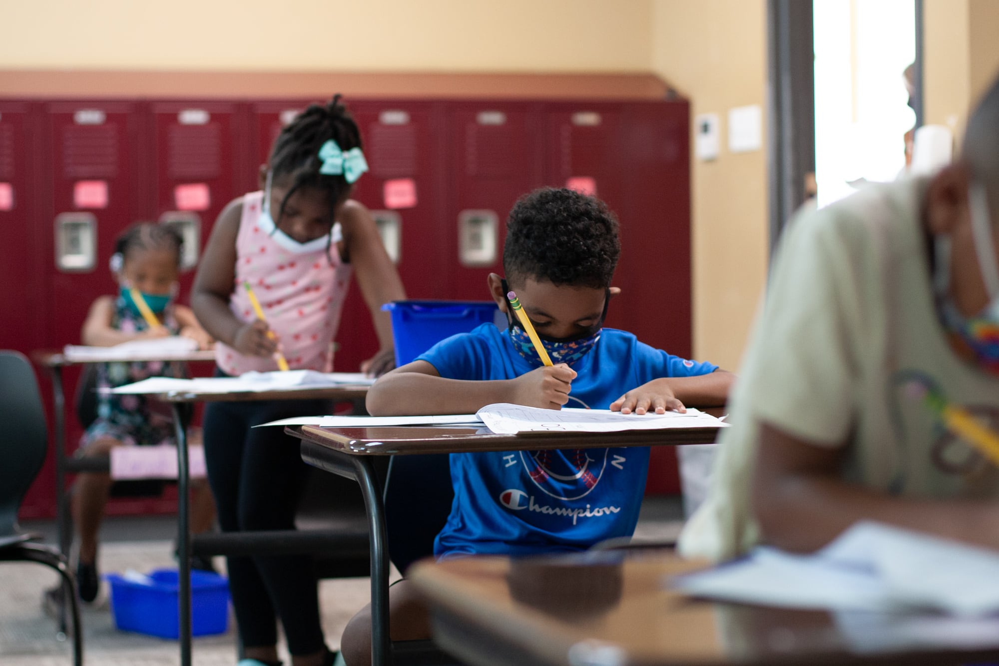 Four students sit in a row of desks in a classroom lined with red lockers. One boy wears a blue shirt and blue mask while writing on a worksheet