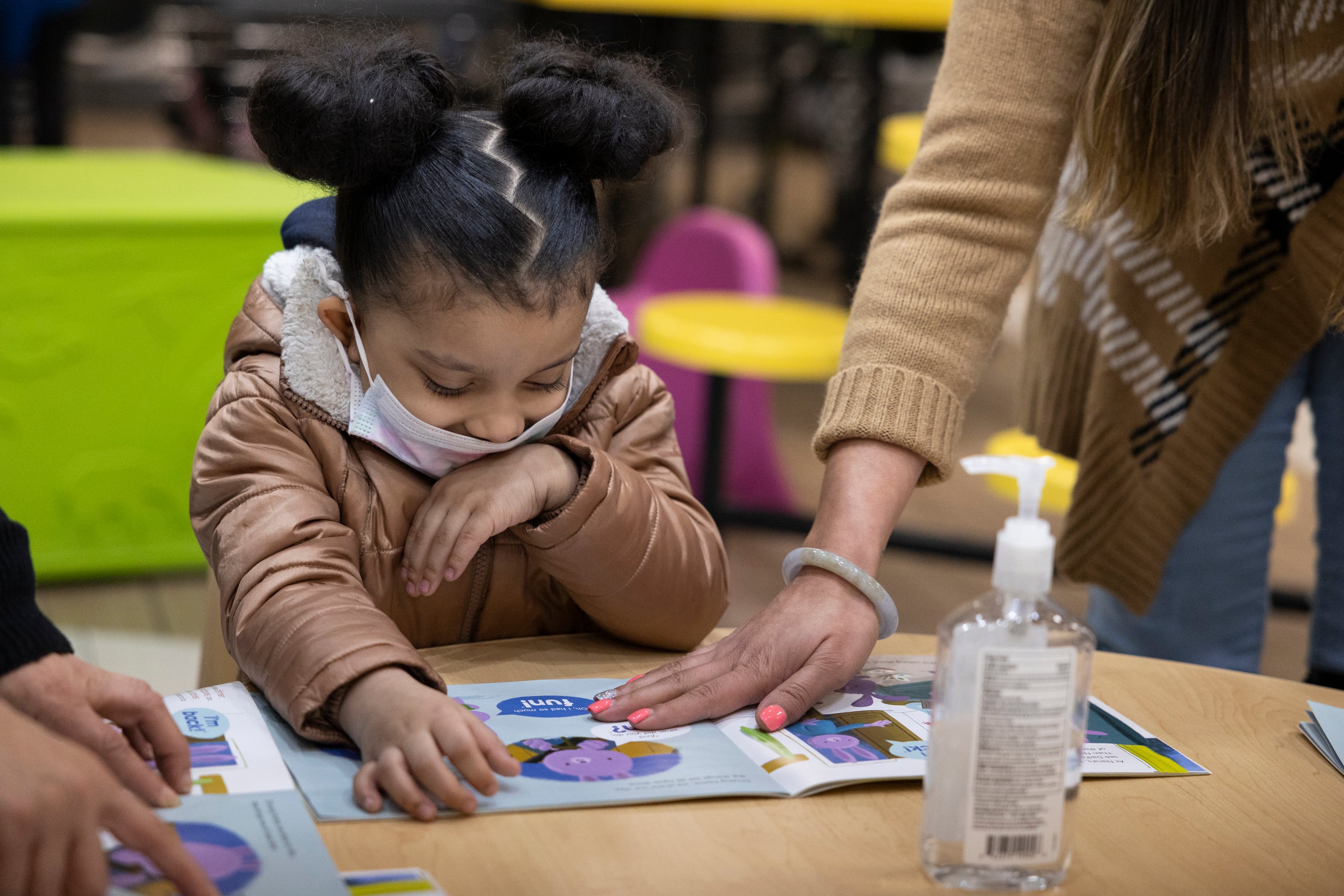 A little girl works on reading a children’s book as a woman with pink nail polish places her hand on the pages.