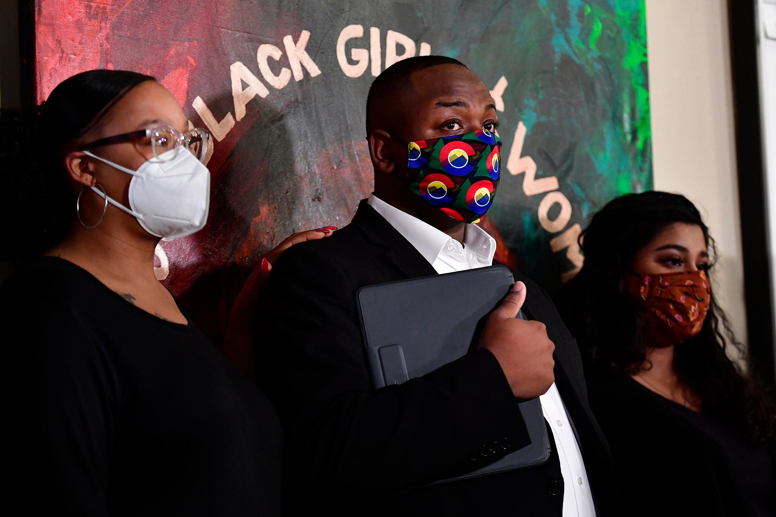 Tay Anderson, wearing a face mask, stands with two women, one on either side of him. A painting on the wall behind him reads “Black Girls and Women.”