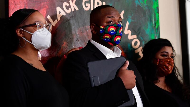 Tay Anderson, wearing a face mask, stands with two women, one on either side of him. A painting on the wall behind him reads “Black Girls and Women.”