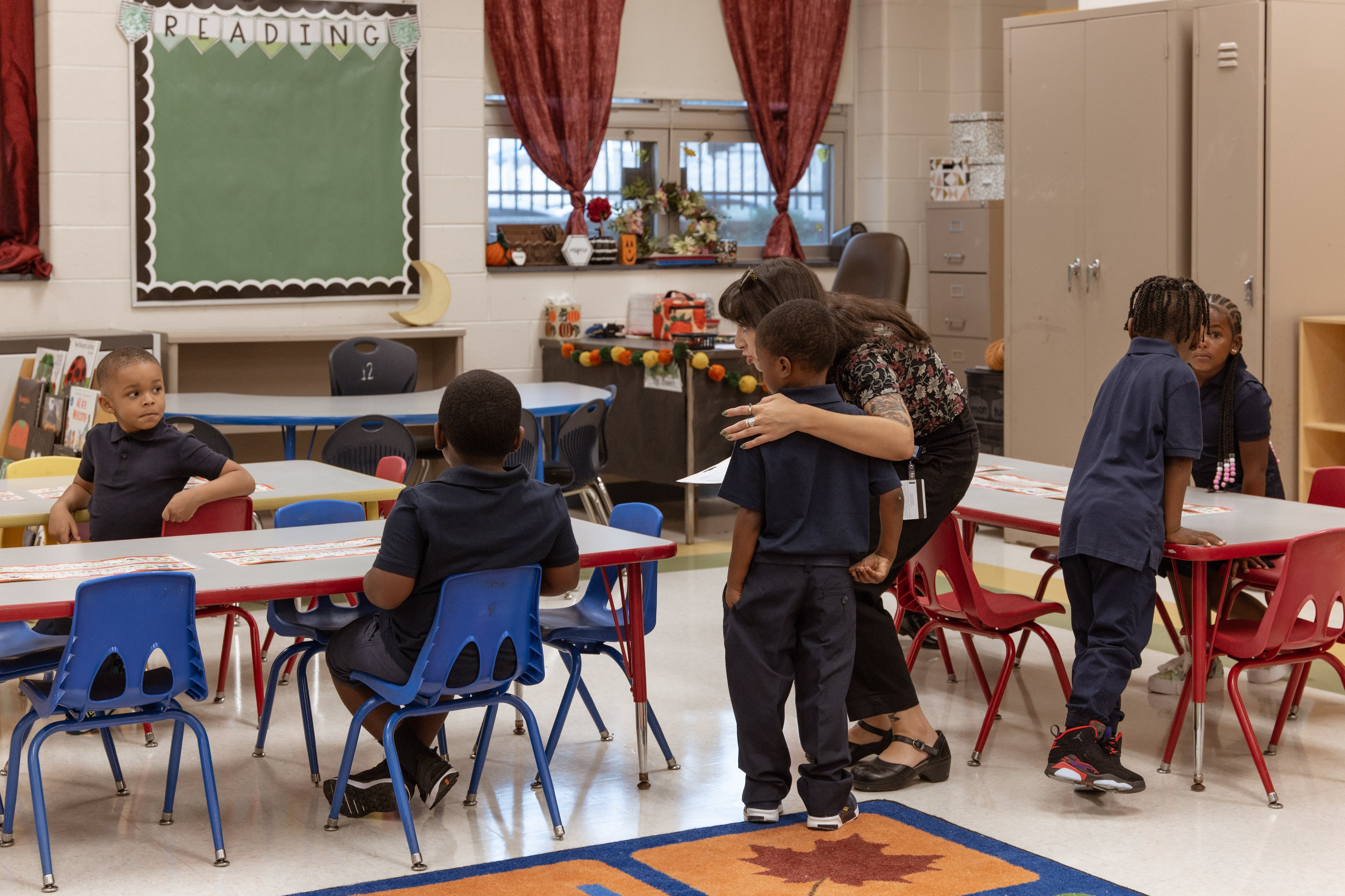 A teacher helps a young student find their seat in a classroom.