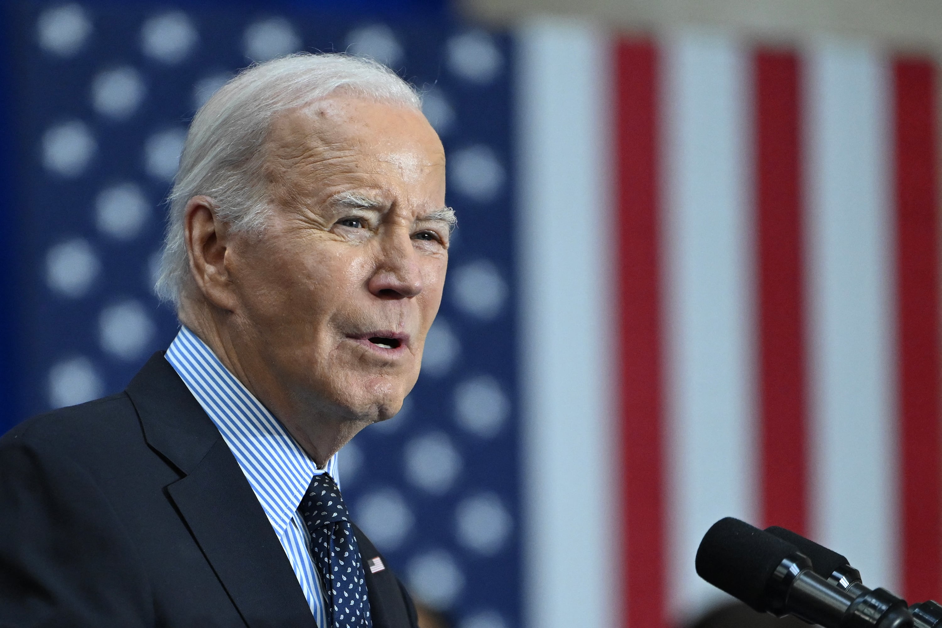 President Biden wears a dark suit and a tie and stands in front of an American flag while speaking at a microphone.