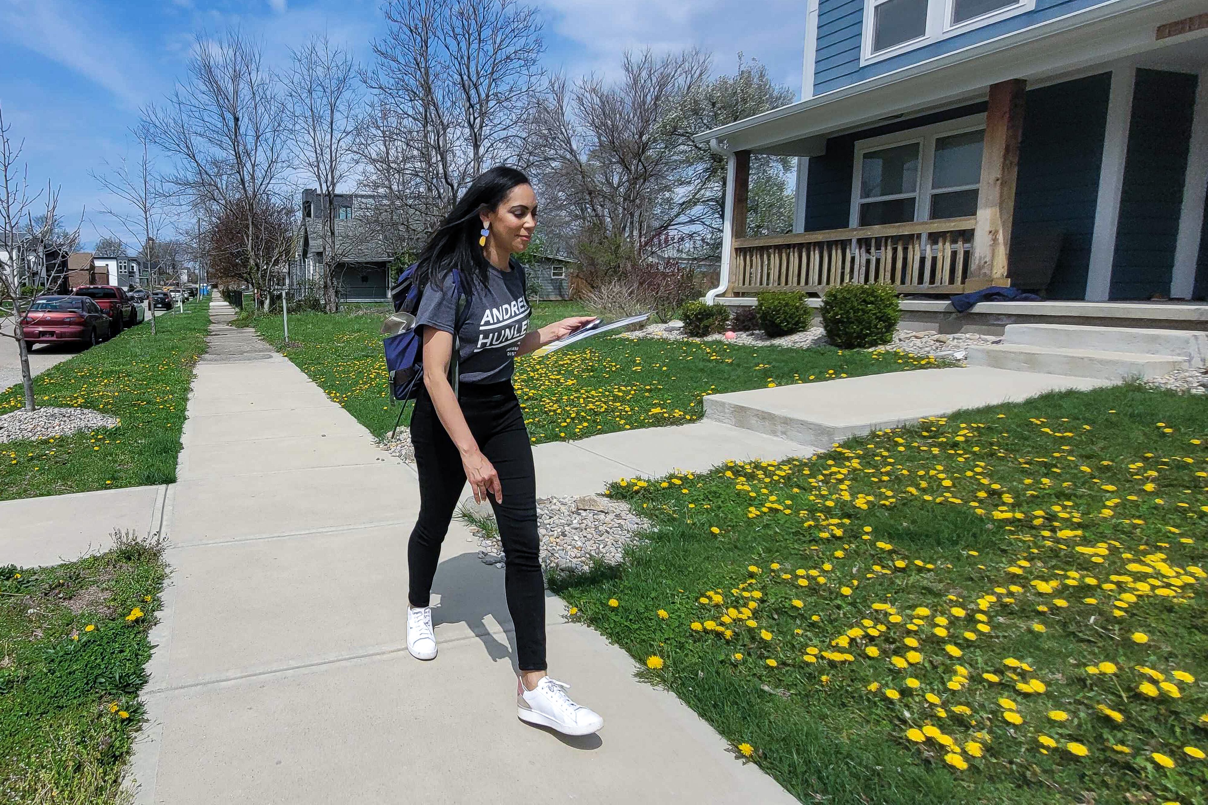 IPS principal Andrea Hunley, wearing a navy blue “Andrea Hunley” T-shirt, black stretch pants, and white athletic shoes, and carrying a clipboard and backpack, walks on a sidewalk past lawns in the Martindale-Brightwood as she campaign for state Senate.
