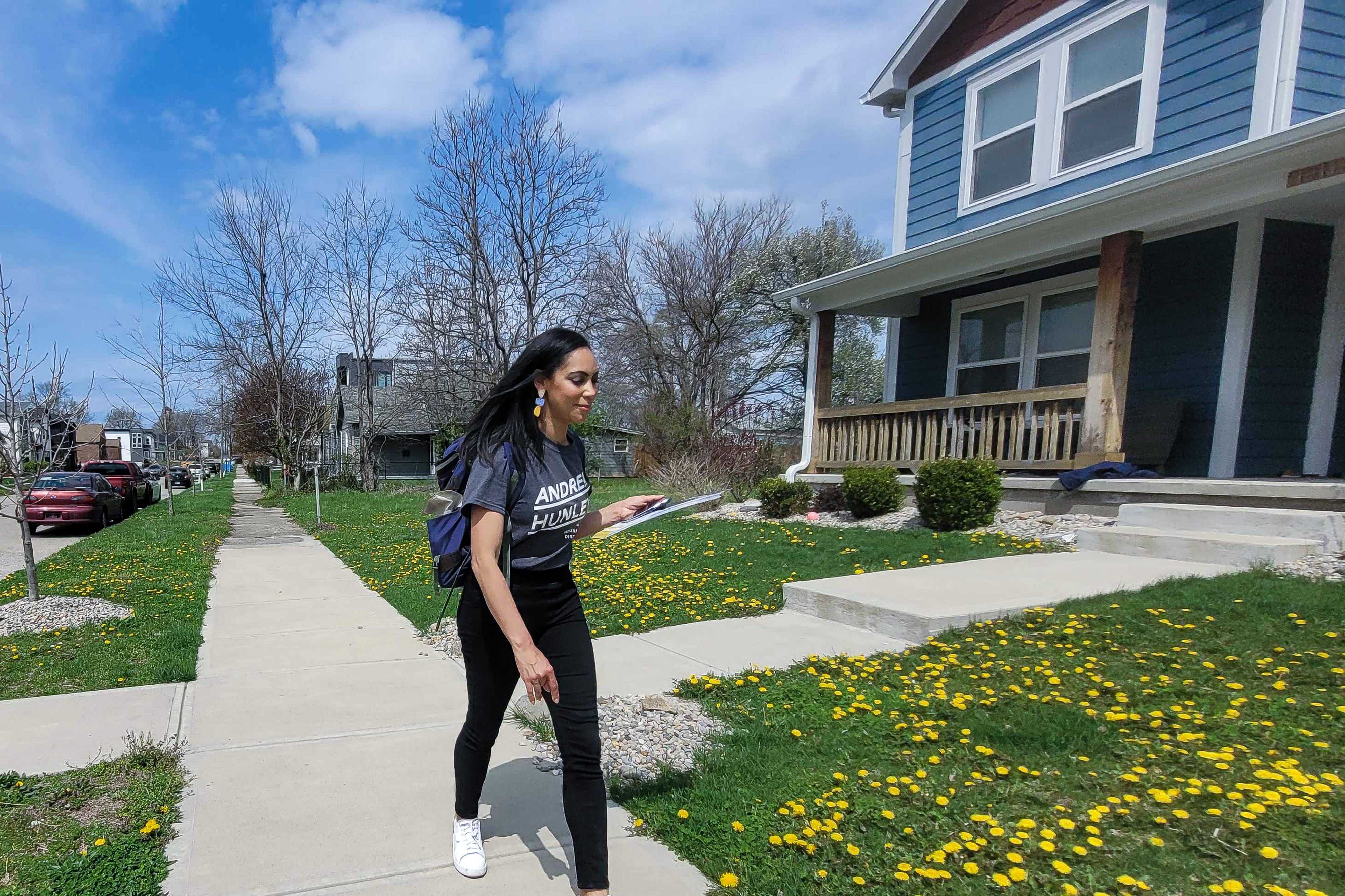 IPS principal Andrea Hunley, wearing a navy blue “Andrea Hunley” T-shirt, black stretch pants, and white athletic shoes, and carrying a clipboard and backpack, walks on a sidewalk past lawns in the Martindale-Brightwood as she campaign for state Senate.