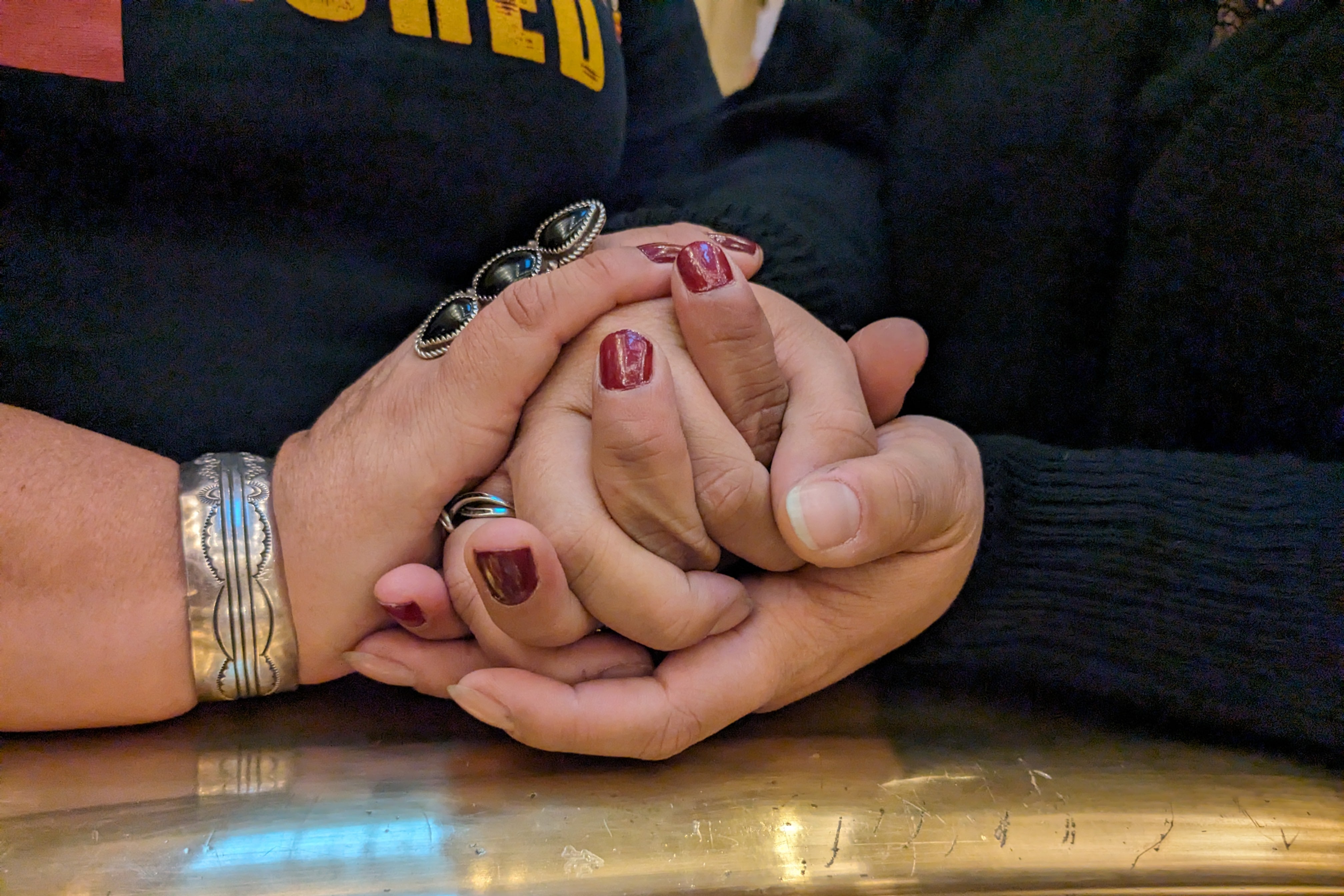 A close up of two people embracing hands. One pair has red nail polish and both people are wearing black shirts.