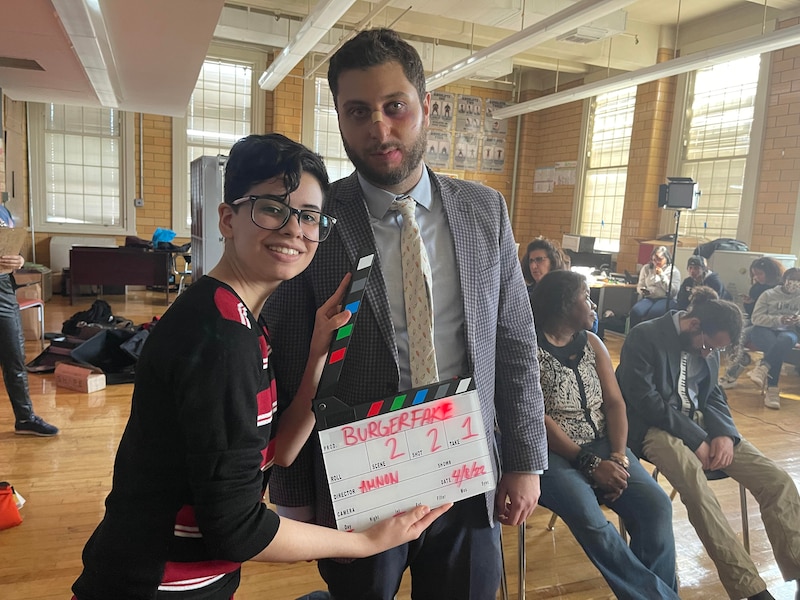 Student with clapperboard stands next to man in a suit