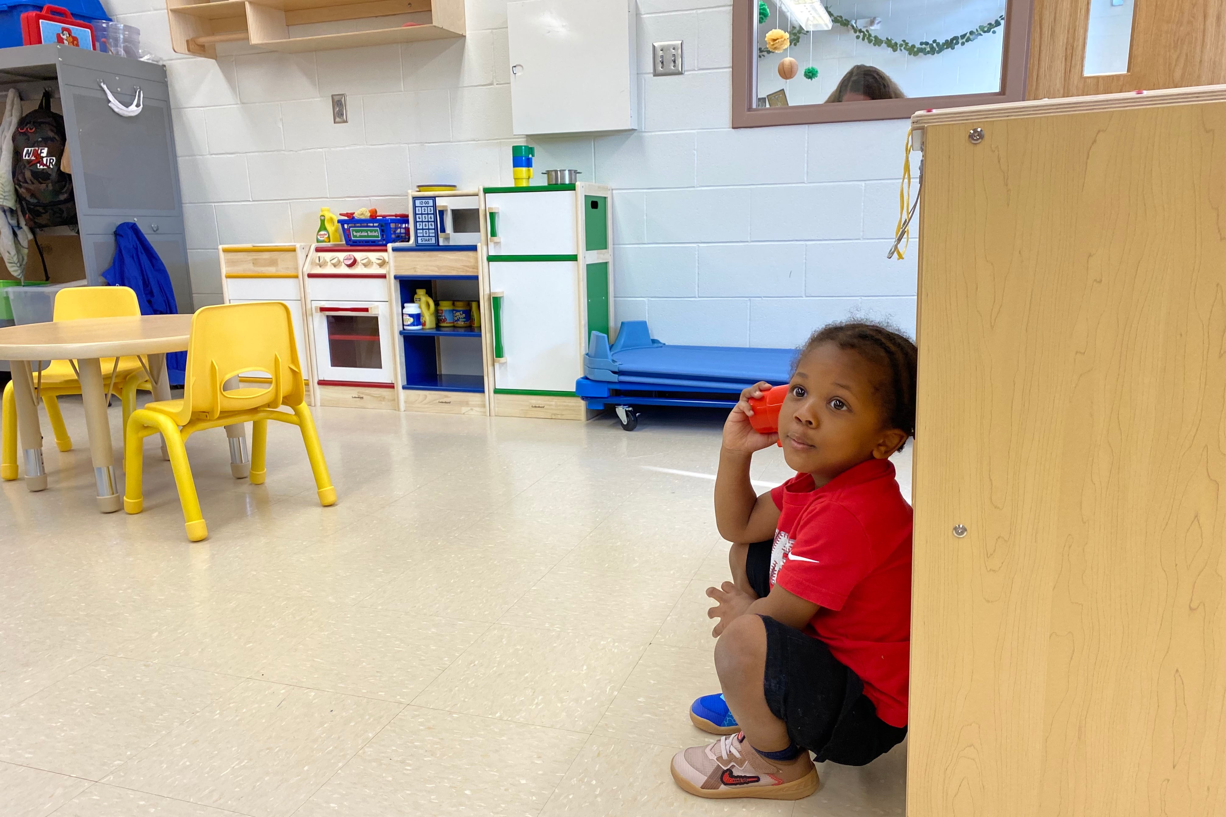 A preschooler wearing a red shirt and two different-colored shoes holds a red cup up to his ear pretending to take a phone order. In the background are a small table and chairs, a play kitchen, and a white board on the wall.
