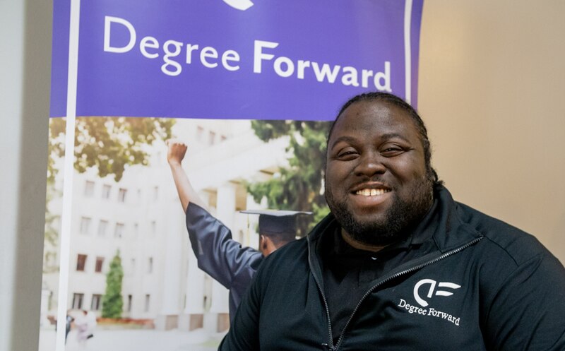 A smiling man wearing a blue jacket with the logo for Degree Forward stands in front of a sign for the program.