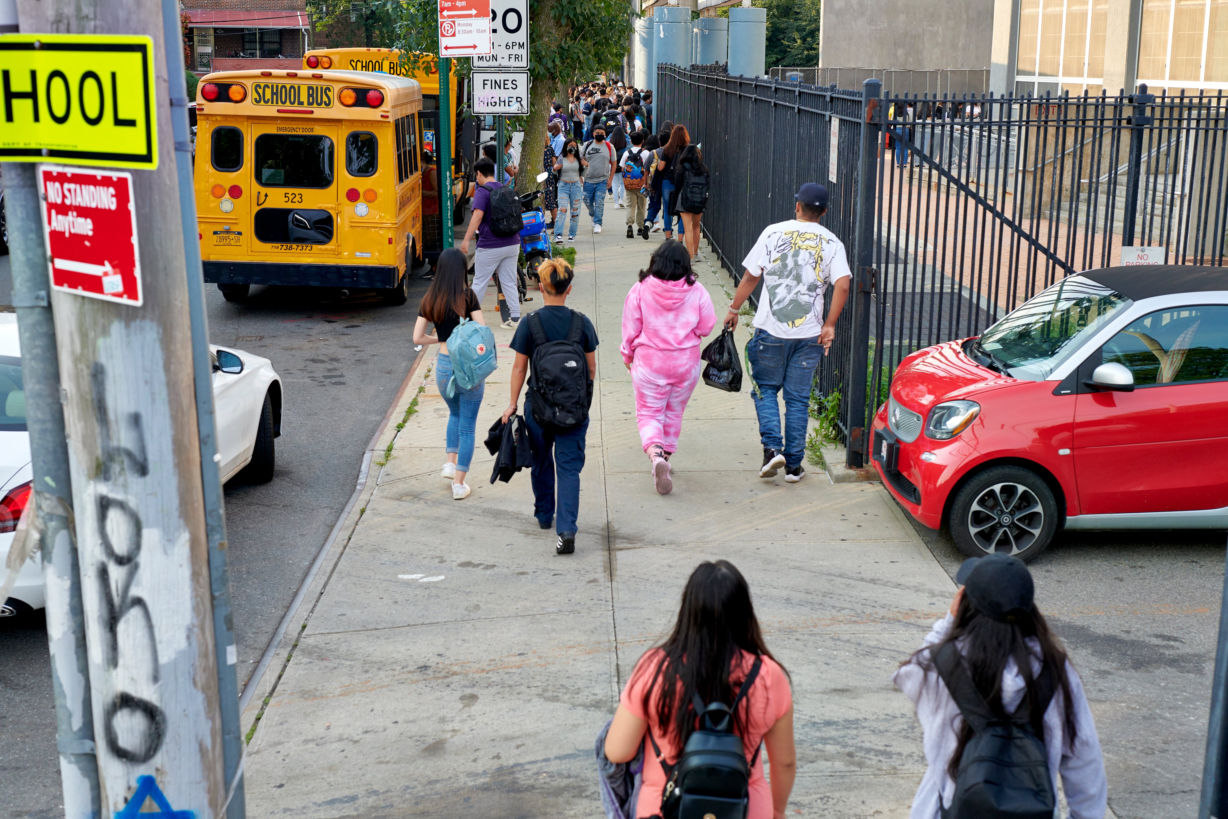 Students, seen from behind, walk on the side walk next to a yellow school bus.