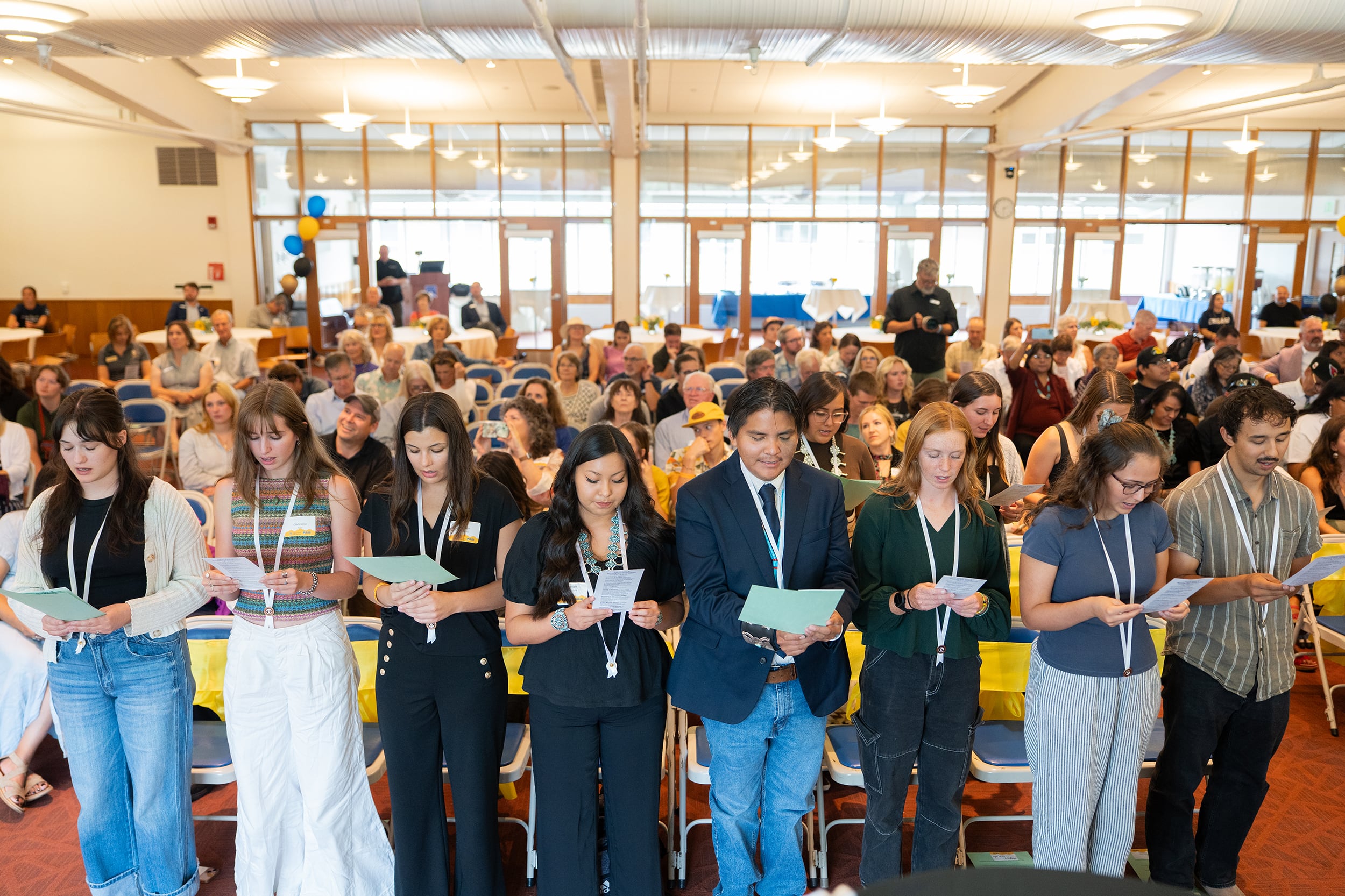 A photograph of a large group of people standing in rows of seats in a large room all reading from a piece of paper.