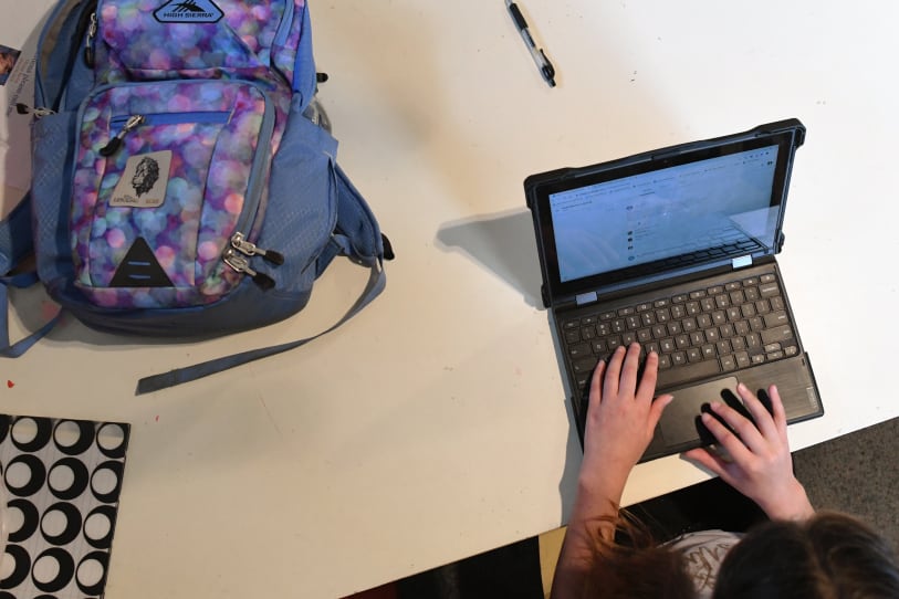 A student works on a laptop at home with her backpack next to her.