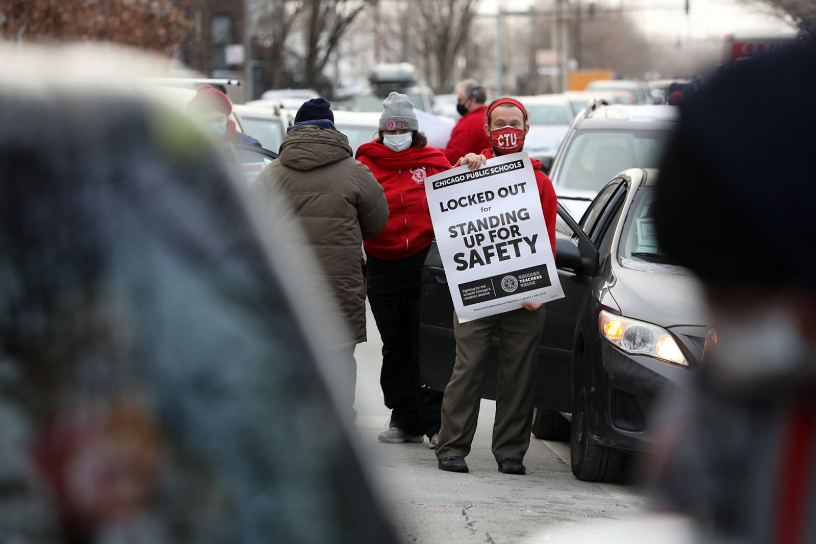 A teacher, wearing a red Chicago Teachers Union mask and sweatshirt, holds a sign that reads, “Locked out for standing up to safety”.