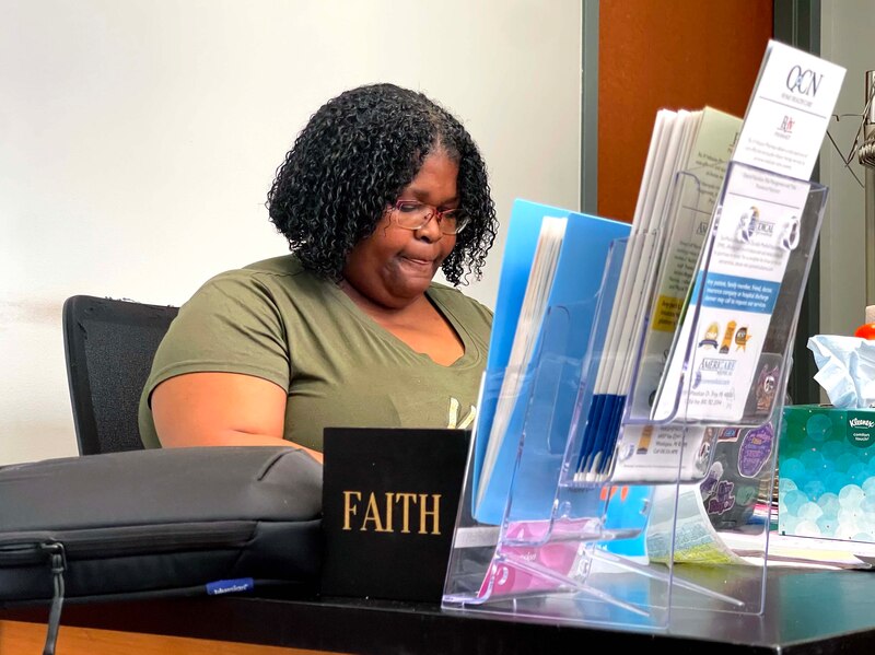 A photograph of a Black woman with short dark, curly hair and wearing a green t-shirt, sits at a desk with pamphlets and other items on her desk in the foreground.
