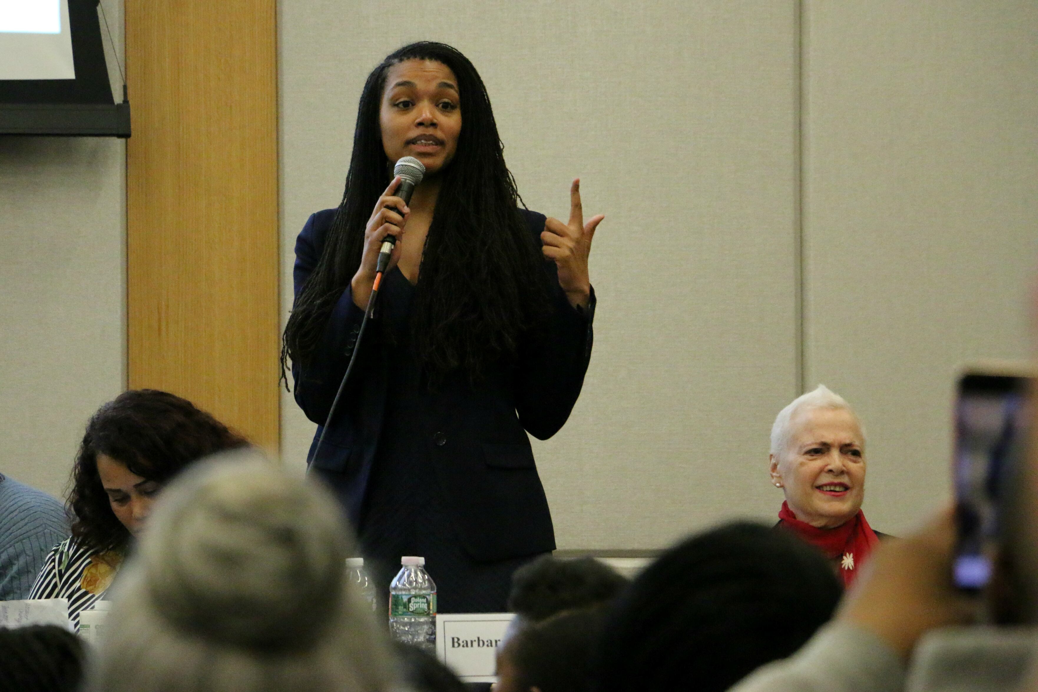 A woman wearing a black blazer holds a microphone as she speaks to a crowd.