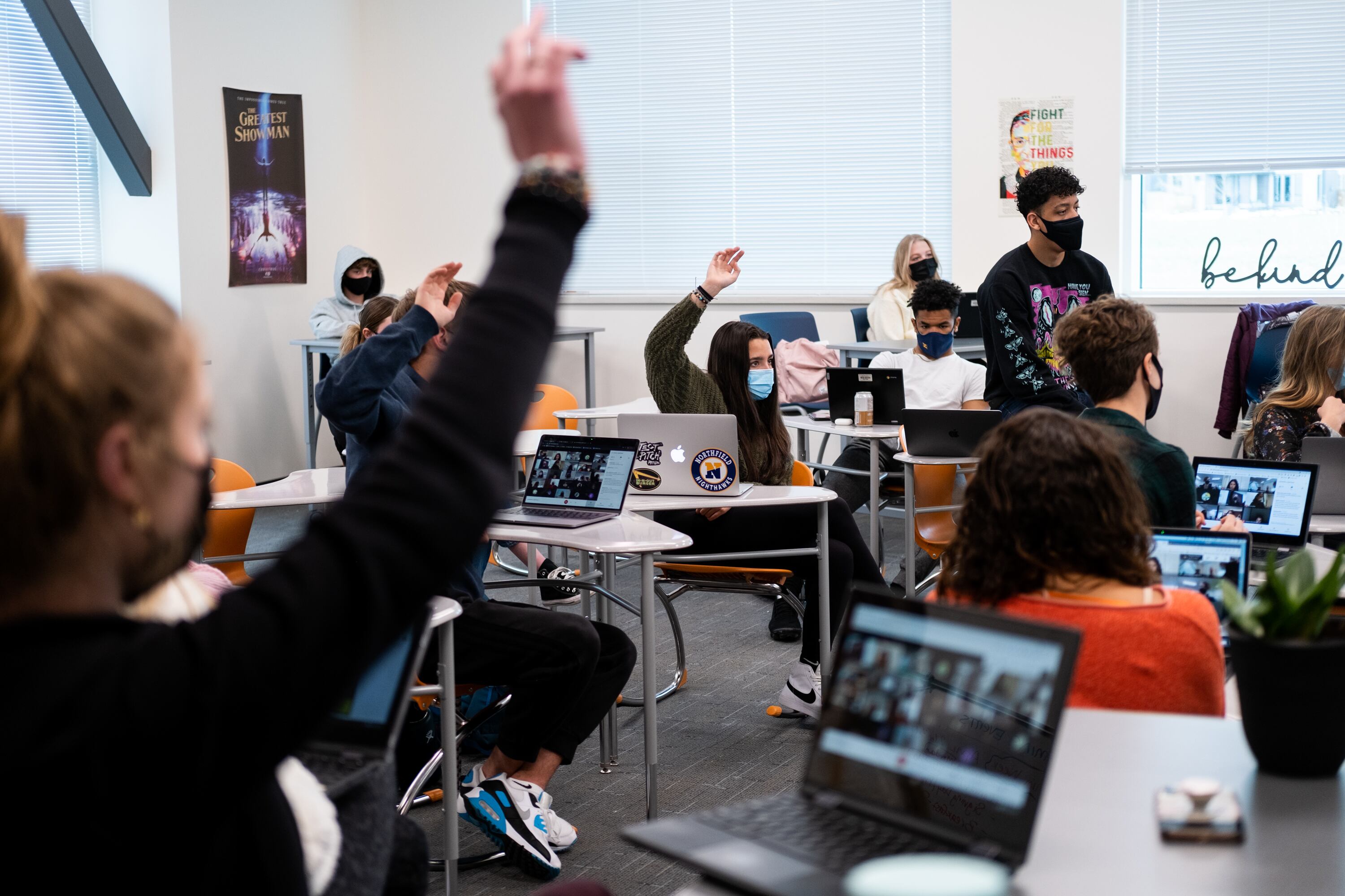 Students wearing masks sit in a Denver high school classroom. The students have open laptops on their desks. Several students are raising their hands.