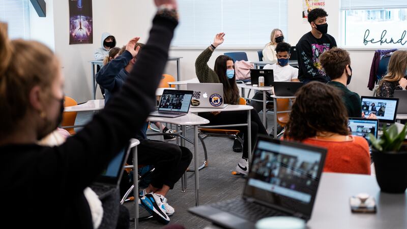 Students wearing masks sit in a Denver high school classroom. The students have open laptops on their desks. Several students are raising their hands.
