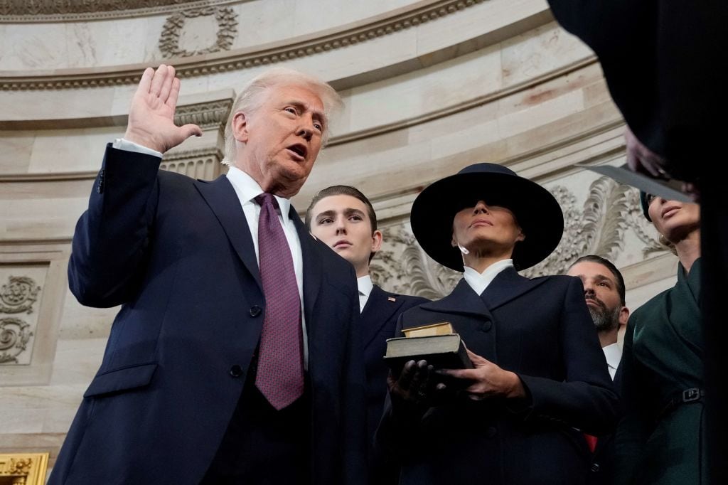Donald Trump, an older man in a dark suit and a maroon tie, raises his right hand to take his oath of office. His wife, Melania Trump, stands by his side wearing a dark suit and dark, white-brimmed hat.