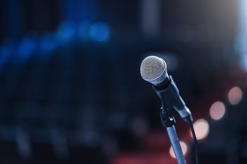 A microphone is in the foreground while a blurred out seating and lights in a concert hall is in the background.