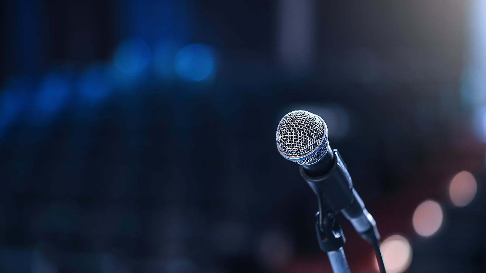 A microphone is in the foreground while a blurred out seating and lights in a concert hall is in the background.