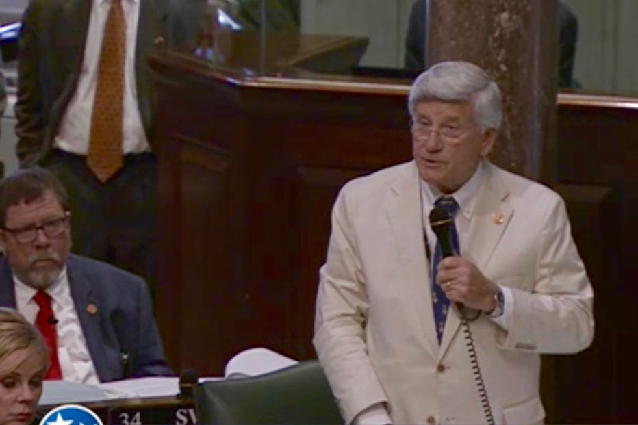 A man in a white suit speaks into a microphone in a legislative chamber.