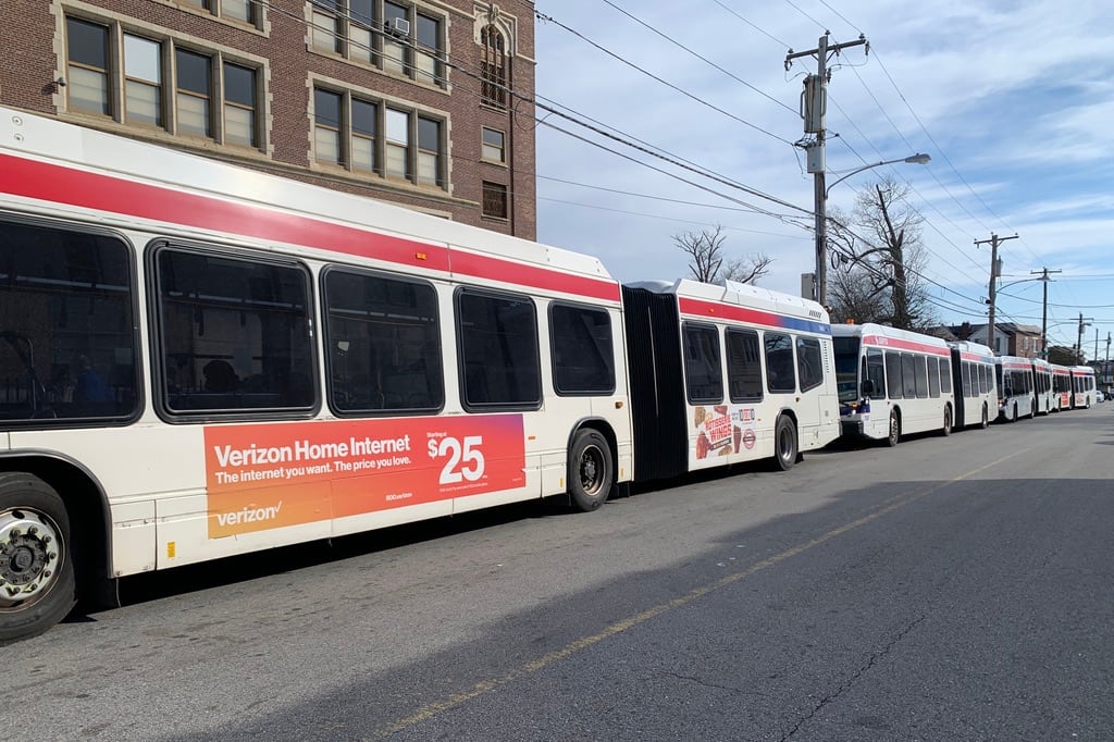 White buses with red strips near the top line up on a street in front of a brick building.