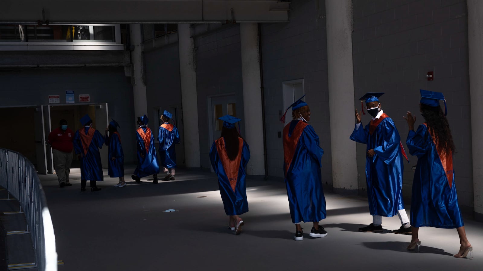 High school seniors in blue and orange caps and gowns walk through the hallway of a stadium to prepare for graduation.