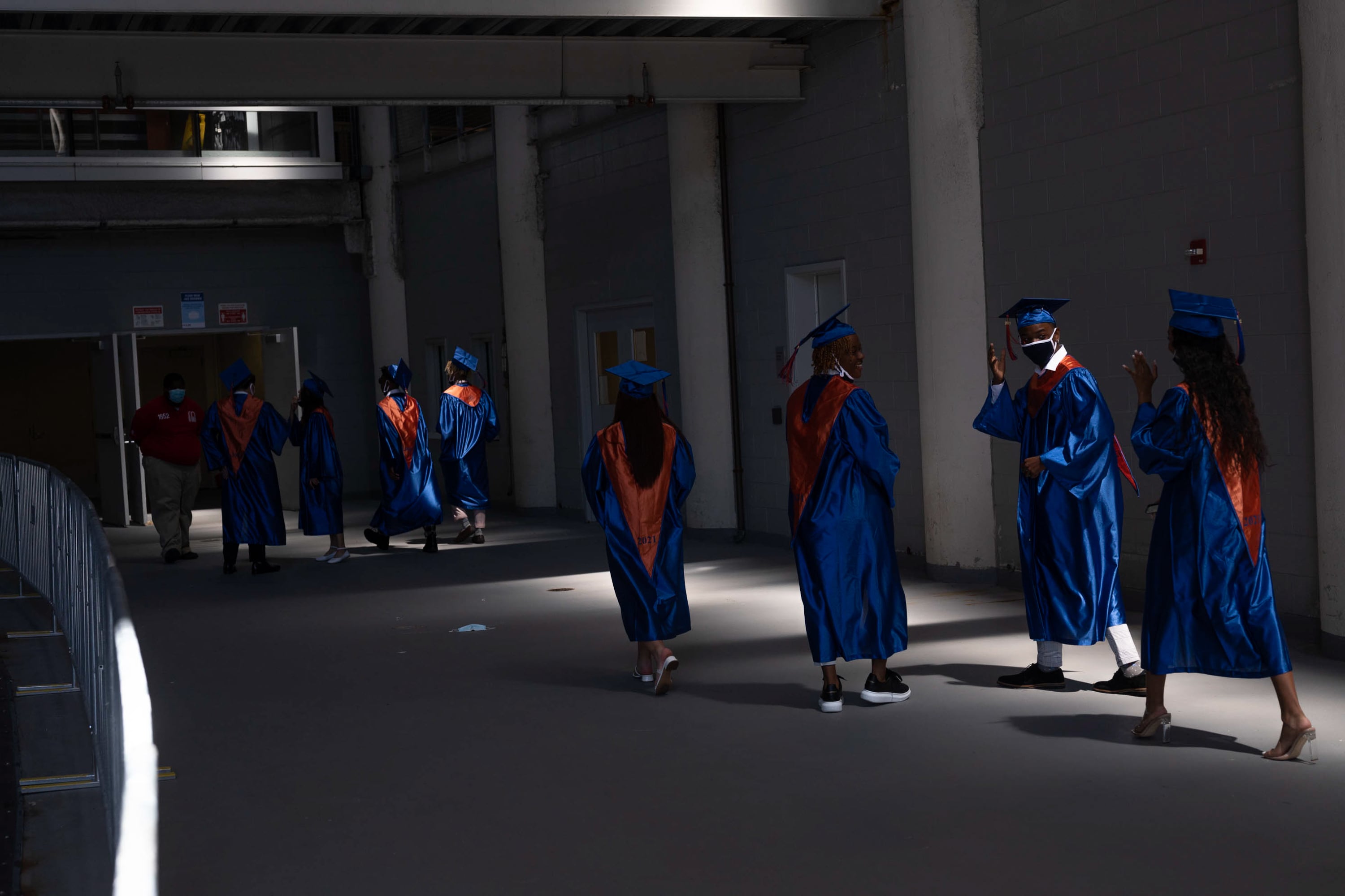 High school seniors in blue and orange caps and gowns walk through the hallway of a stadium to prepare for graduation.