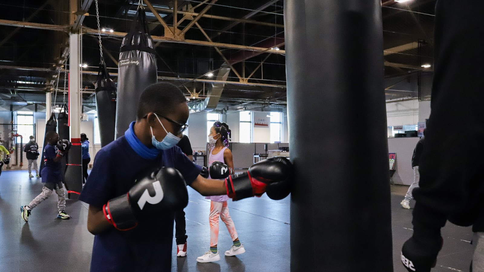 A boy hits a heavy bag as other young students also work on their boxing in a large gym.
