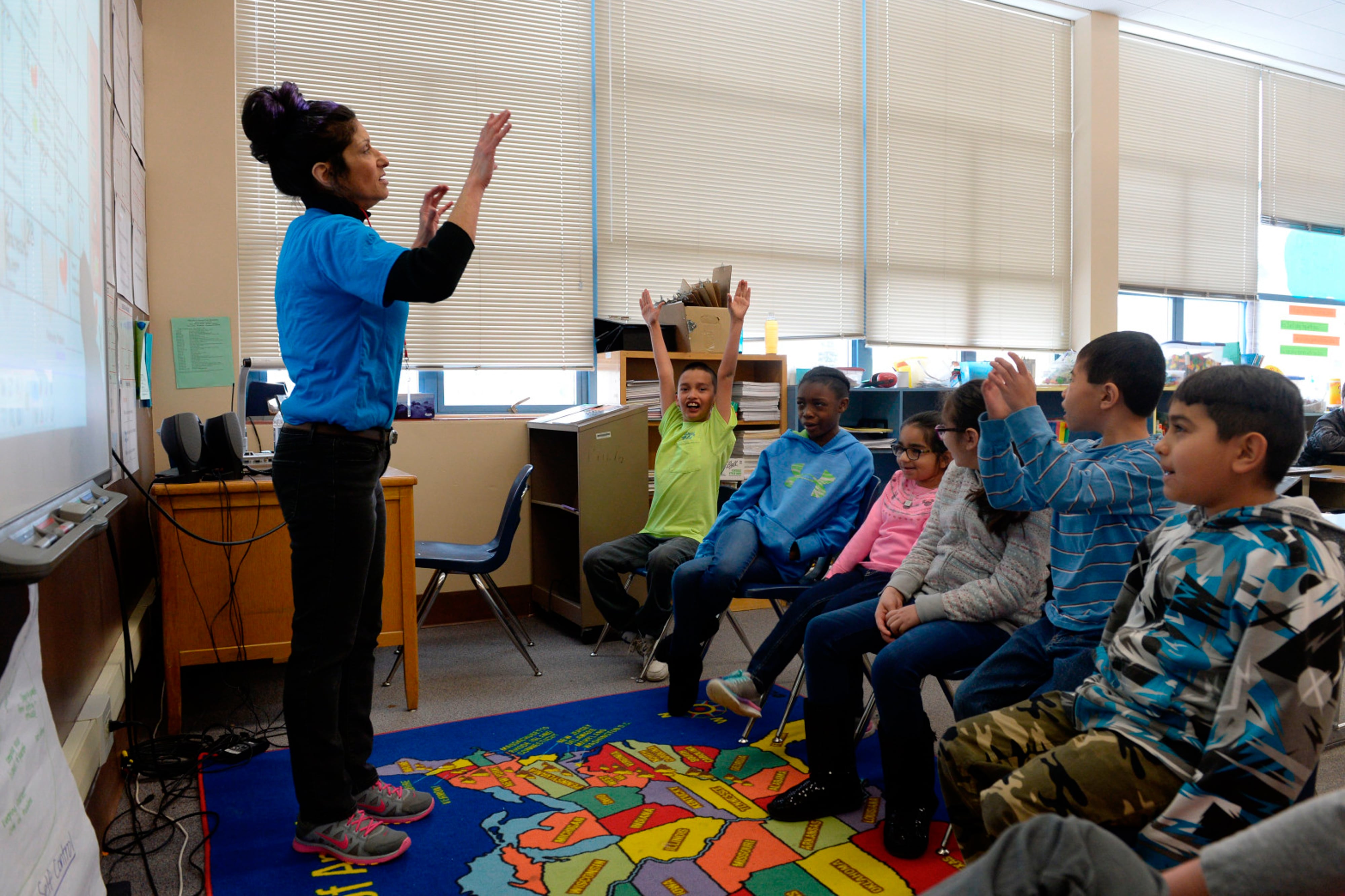 A group of six elementary students sit together and engage with their teacher, standing at the front of the classroom with her arms raised.