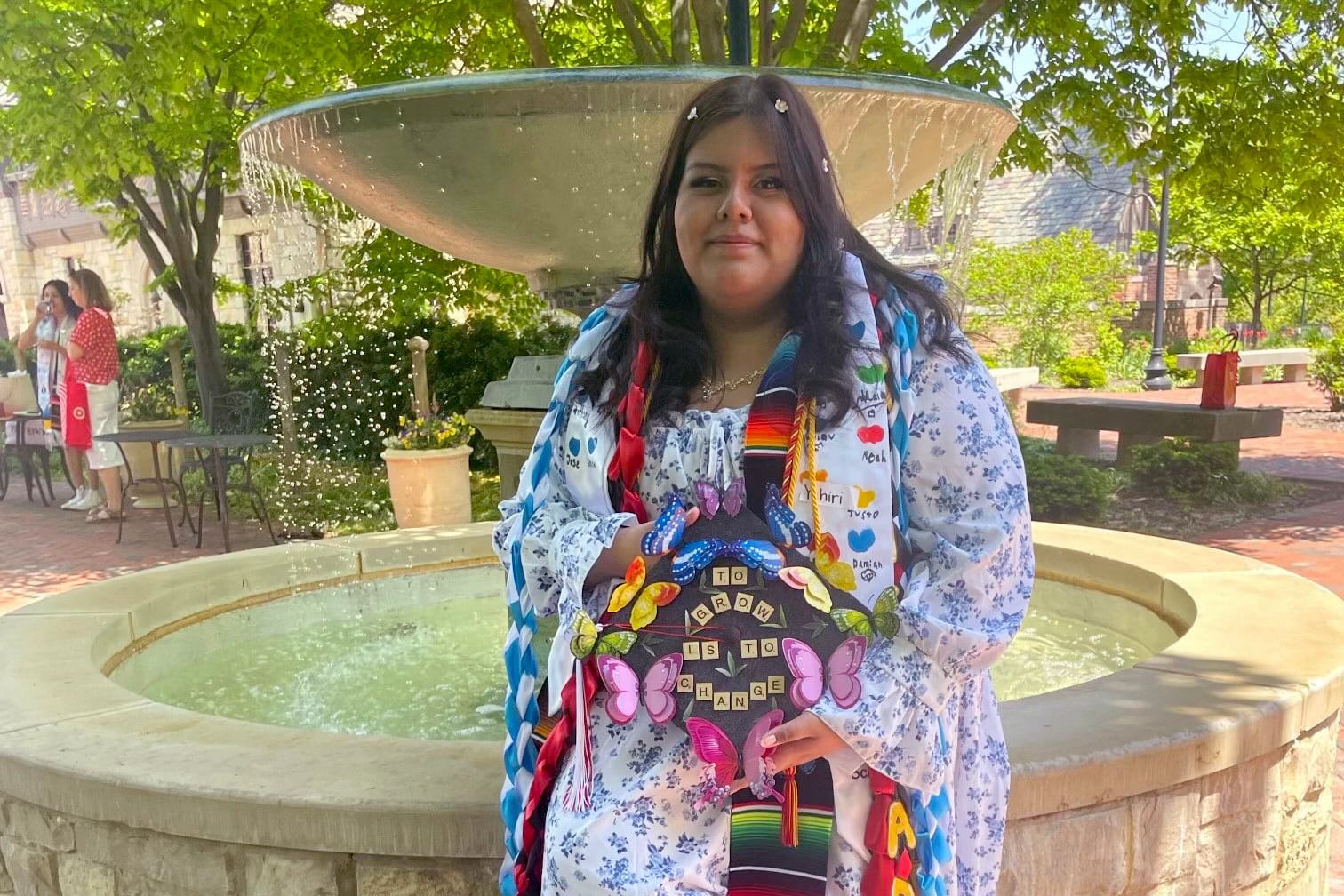 A photograph of a woman with medium length dark hair wearing a floral dress and holding her graduation cap sitting at a fountain outside.