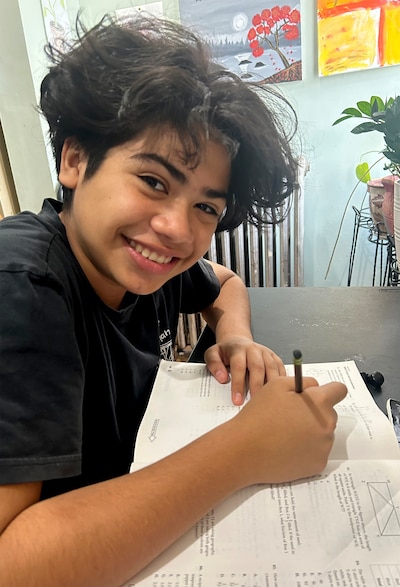 A young boy holds a pencil while smiling and posing for a photo. Theres a school work book on the table and various art pieces on the wall in the background.