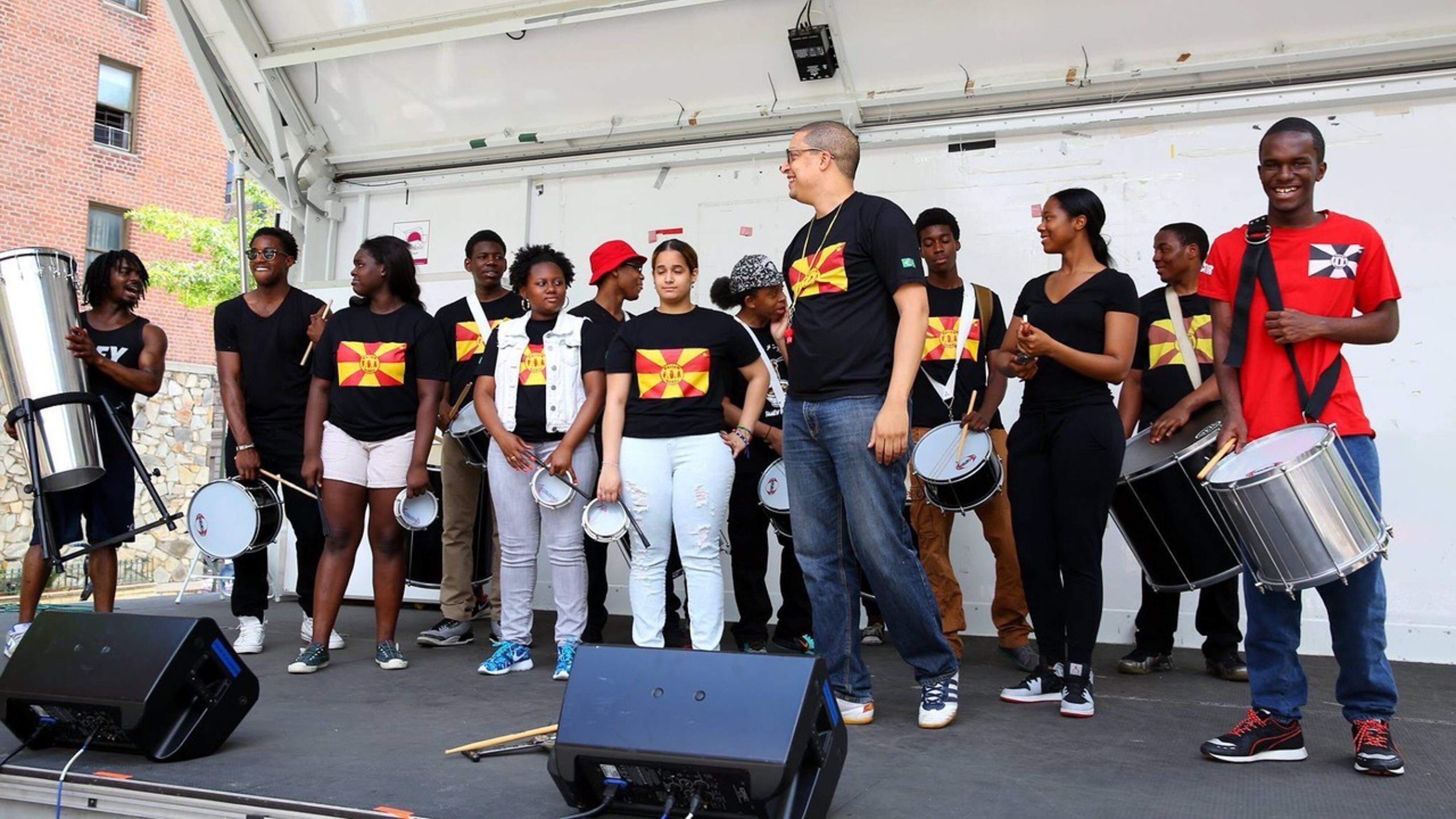 Students and members of Harlem Samba stand with their percussion instruments on a stage, most of them wearing black shirts with red and yellow flags.