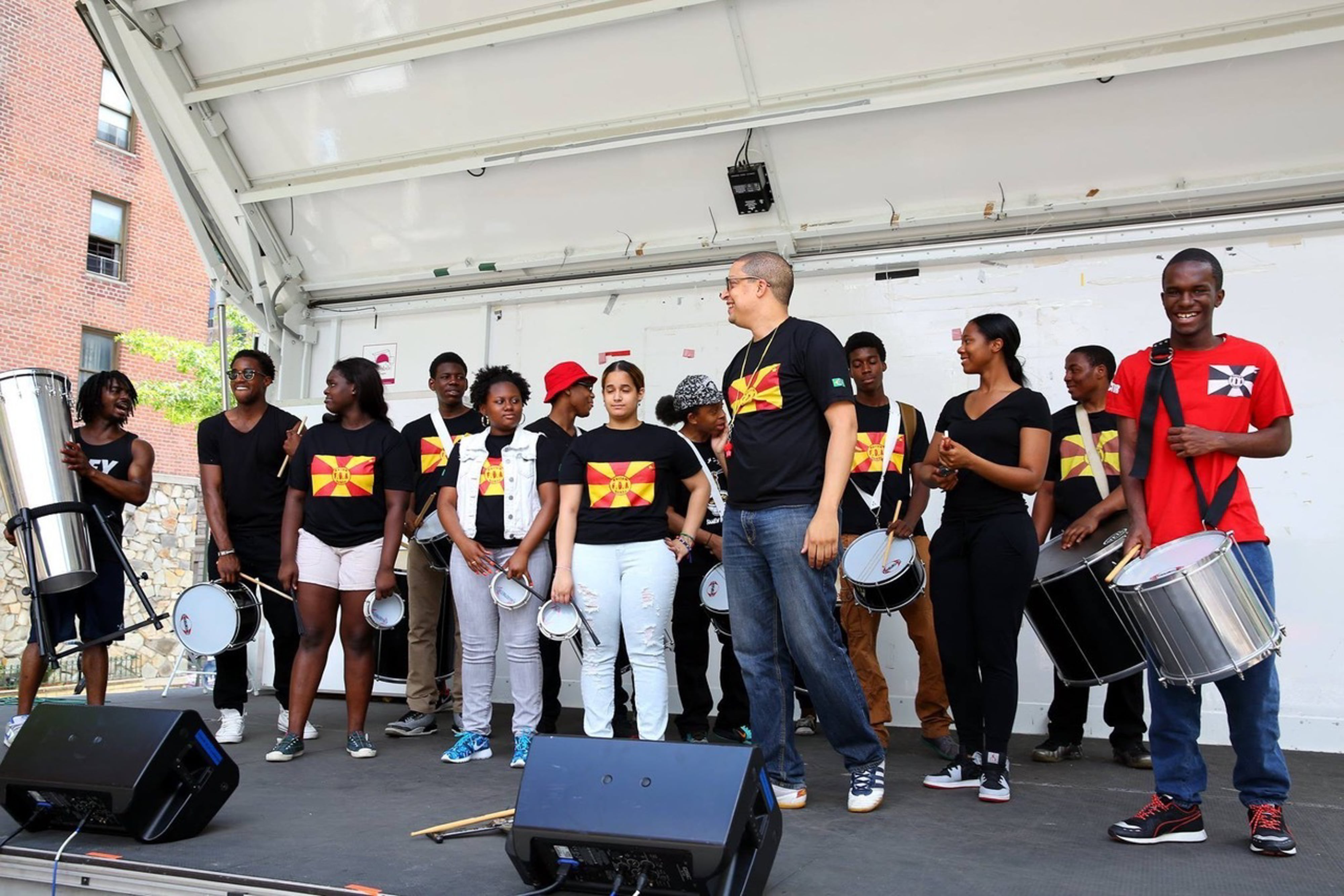 Students and members of Harlem Samba stand with their percussion instruments on a stage, most of them wearing black shirts with red and yellow flags.