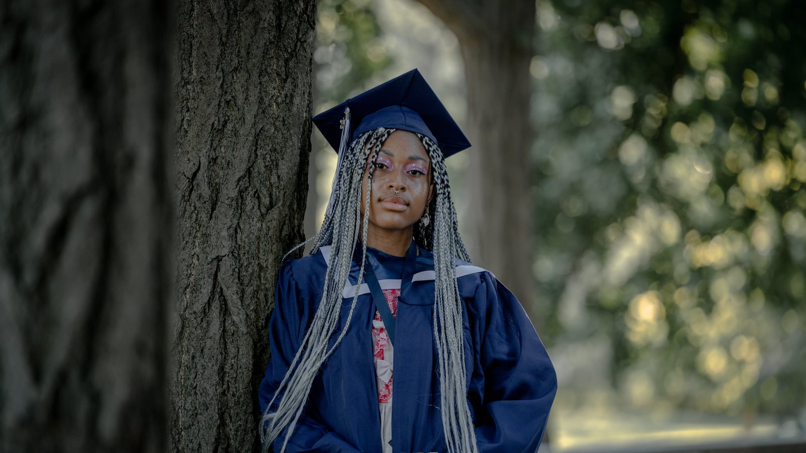 A young woman with long, braided hair poses for a portrait on her graduation day, wearing a blue cap and gown.