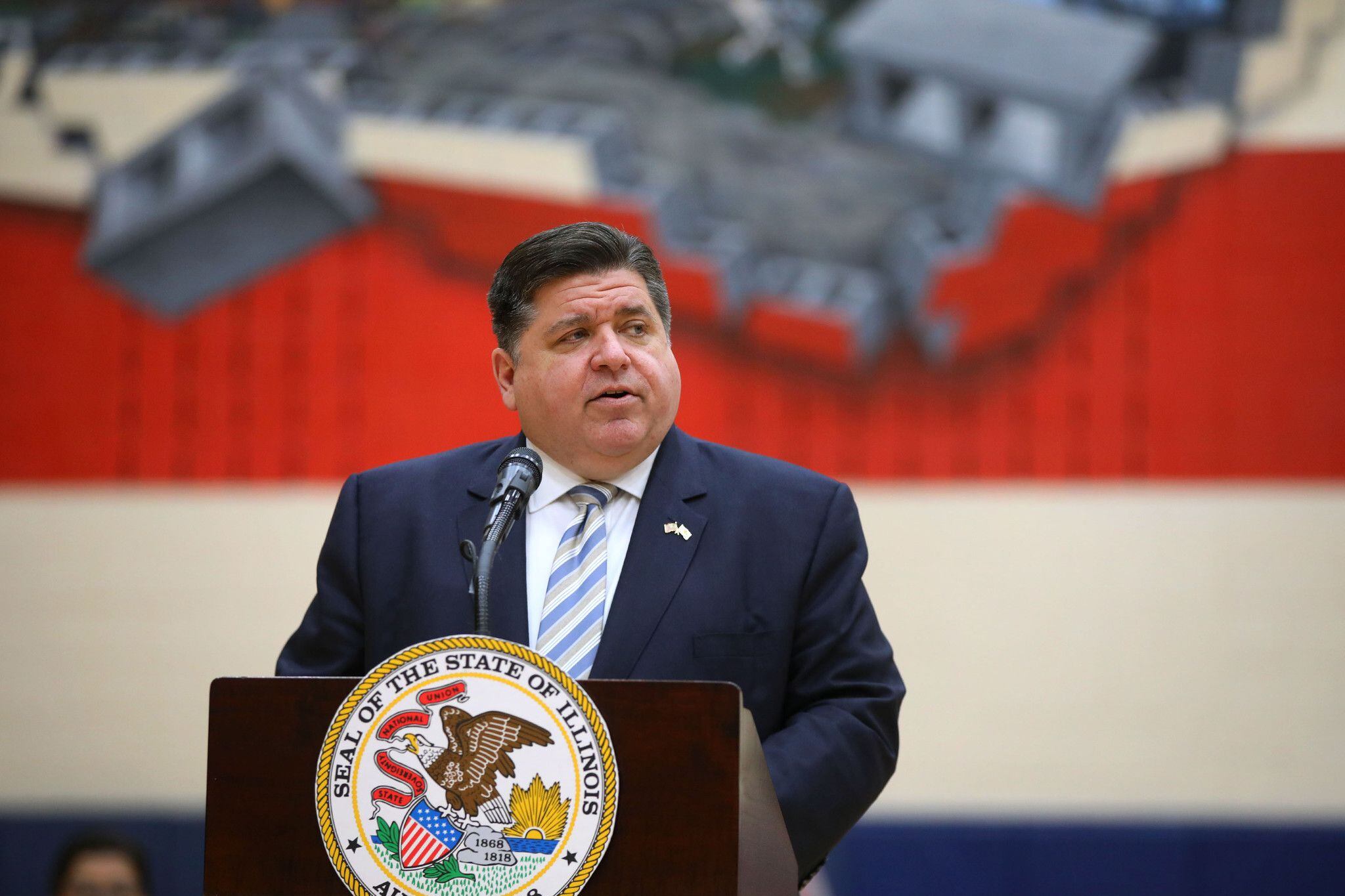 Illinois Governor J.B. Pritzker speaks at a podium in front of a large mural of the Illinois state flag.
