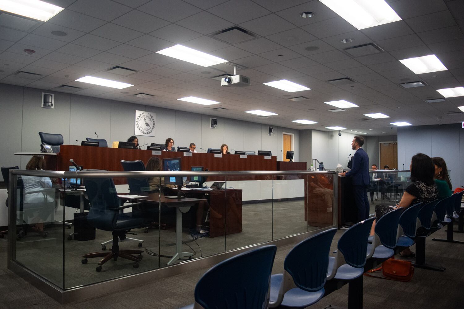 Four members of the Chicago school board sits at an elevated, shared wooden dais, about six feet from a clear glass barrier. Against the barrier is a lectern, where a man speaks into a microphone, facing the school board. The barrier does not reach floor to ceiling. It stops so that people at the lectern can have their heads over it. Behind the man speaking is a row of chairs, where two audience members are in the frame.