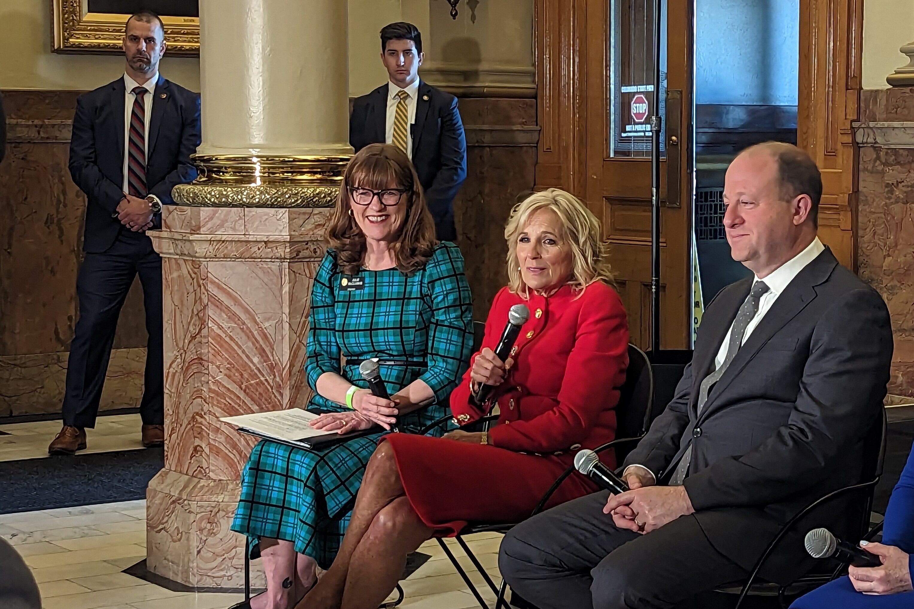 First lady Jill Biden dressed in red sits with Colorado’s House speaker dressed in teal and Gov. Jared Polis.