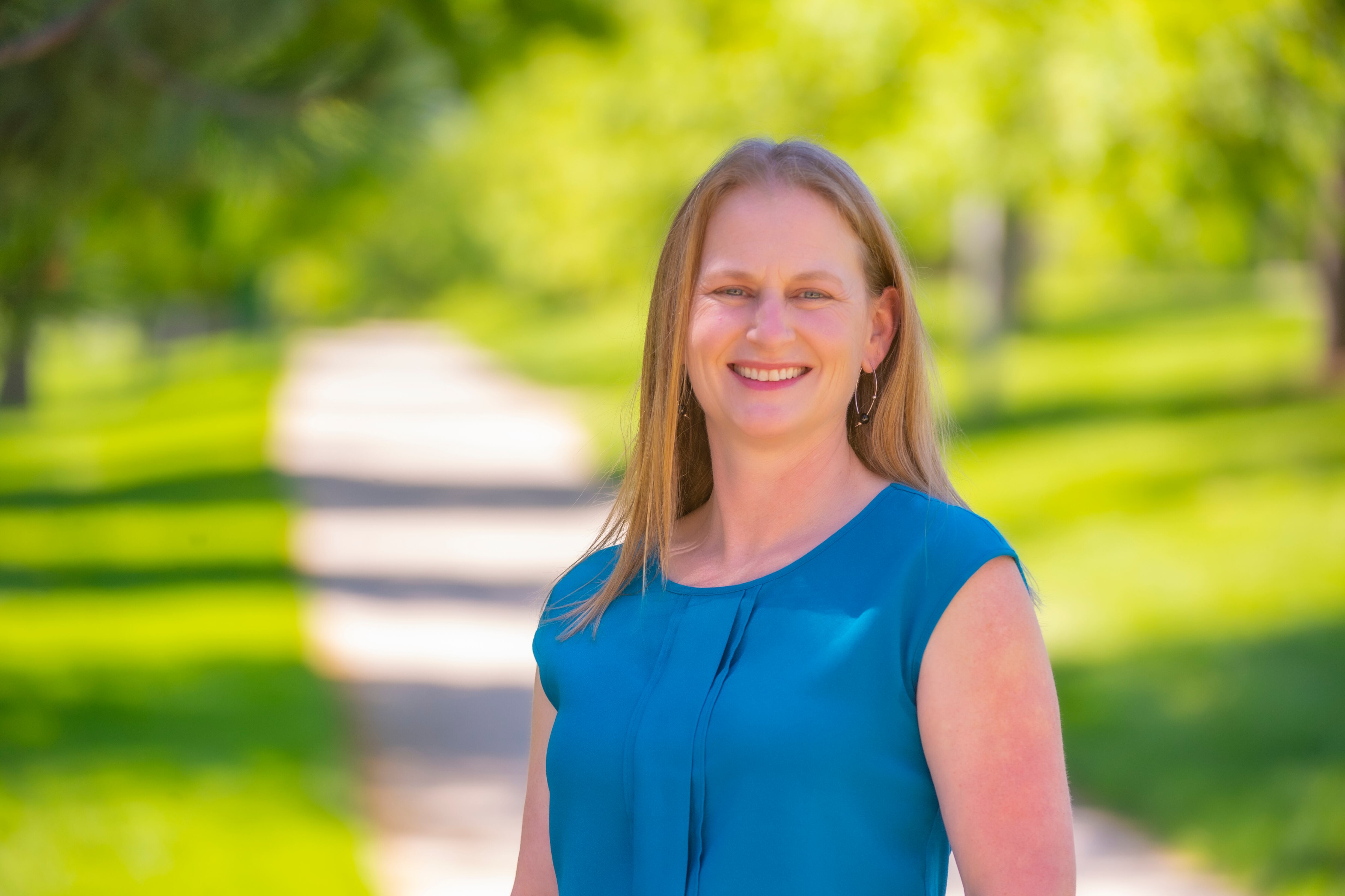 A blonde woman in a blue shirt smiles at the camera. She is standing outside.