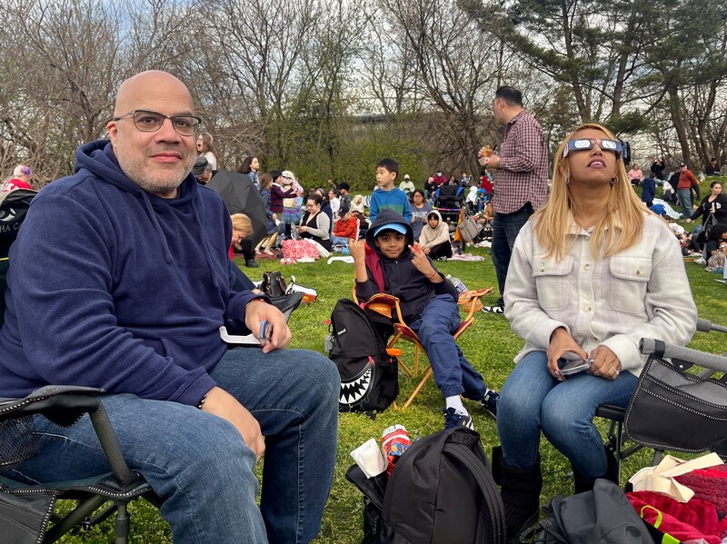 Two adults and one child sit on lawn chairs next to each other posing for a photograph with a large group of people in the background.