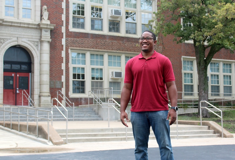 A man wearing a red shirt poses for a portrait outside the front entrance of a school building.