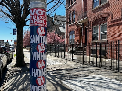 A street pole covered in candidate signs on a sidewalk in front of a red brick building.